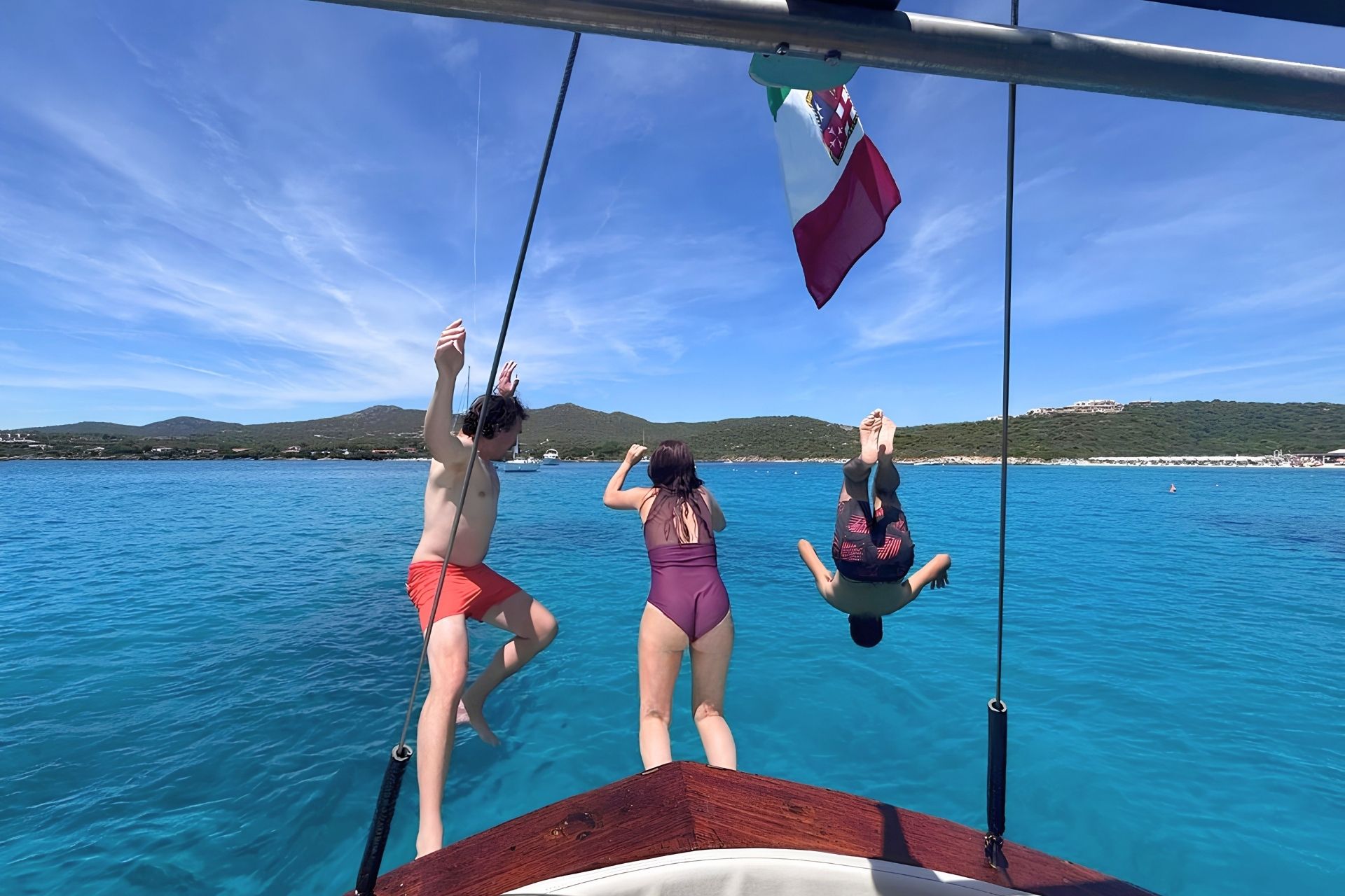 Trois personnes s'amusent sur un voilier d'époque, avec une île en arrière-plan et une île plus éloignée au loin. Le bateau est amarré et bat pavillon italien.