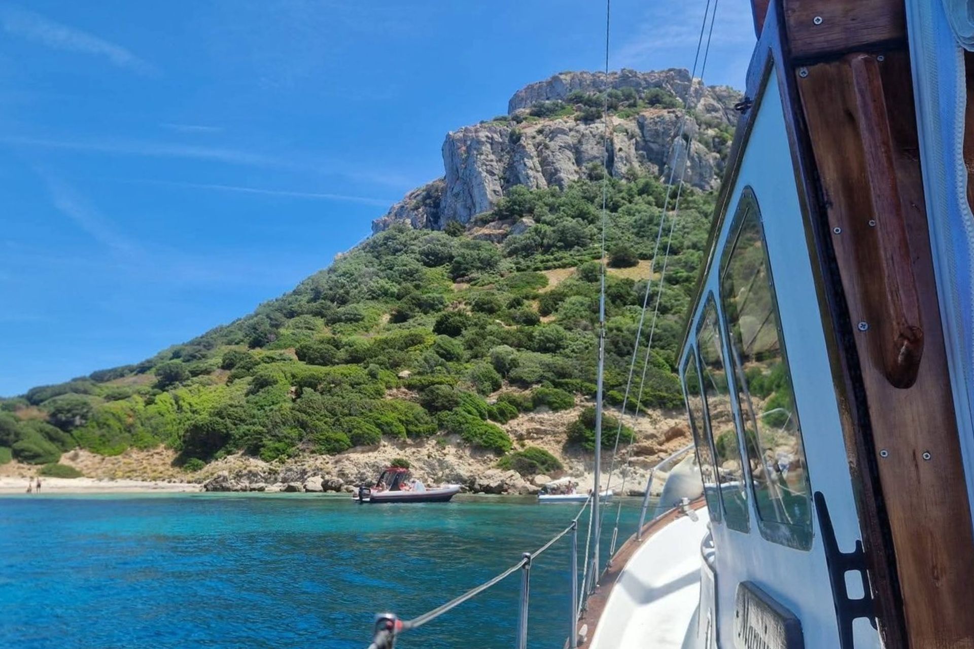 Un voilier d'époque navigue dans des eaux cristallines près d'une côte rocheuse verdoyante. Un autre bateau plus petit se trouve plus loin.