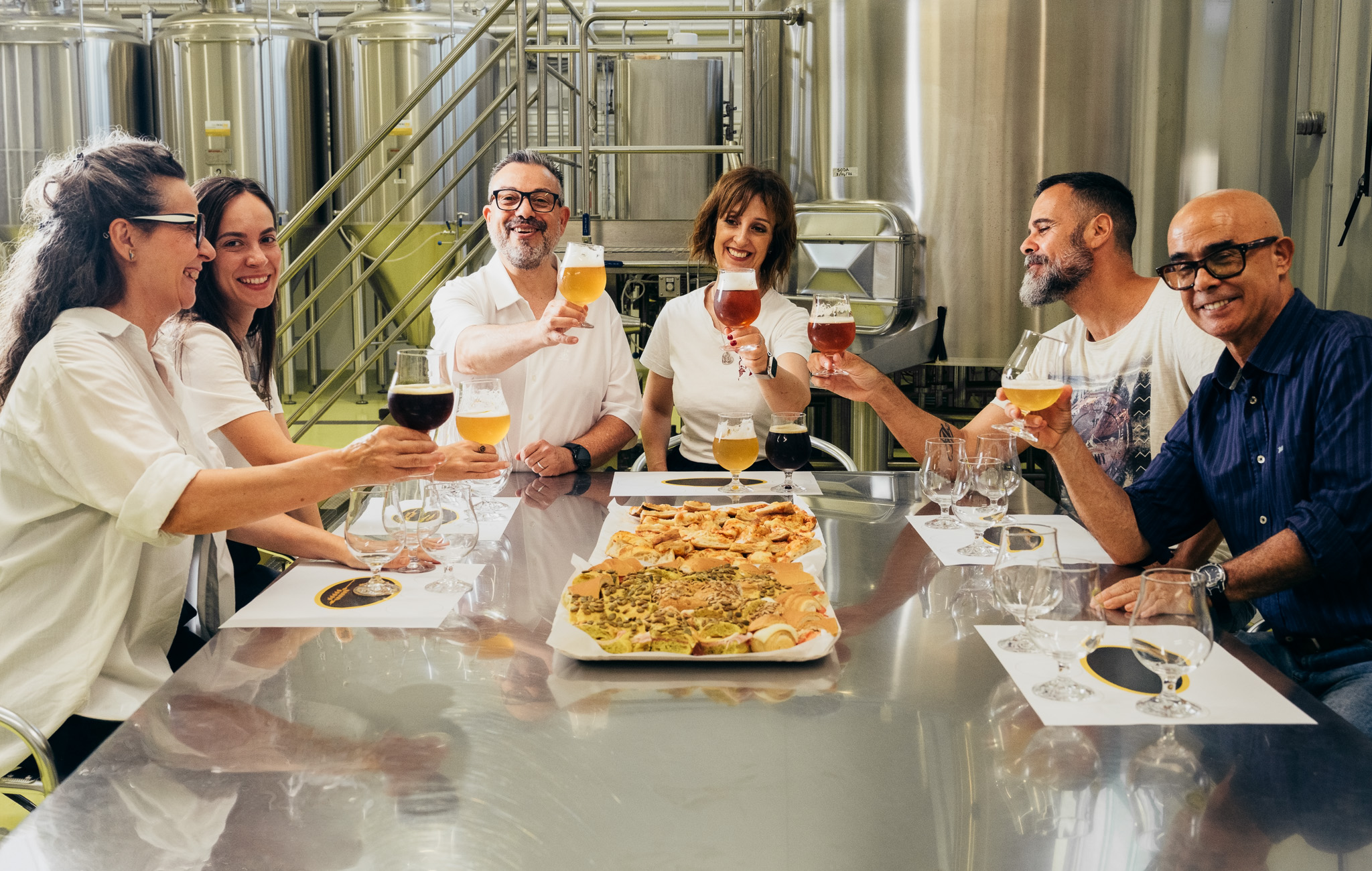 Gruppo di persone che brinda con bicchieri di birra artigianale in un birrificio, con sfondo di serbatoi e attrezzature industriali.
