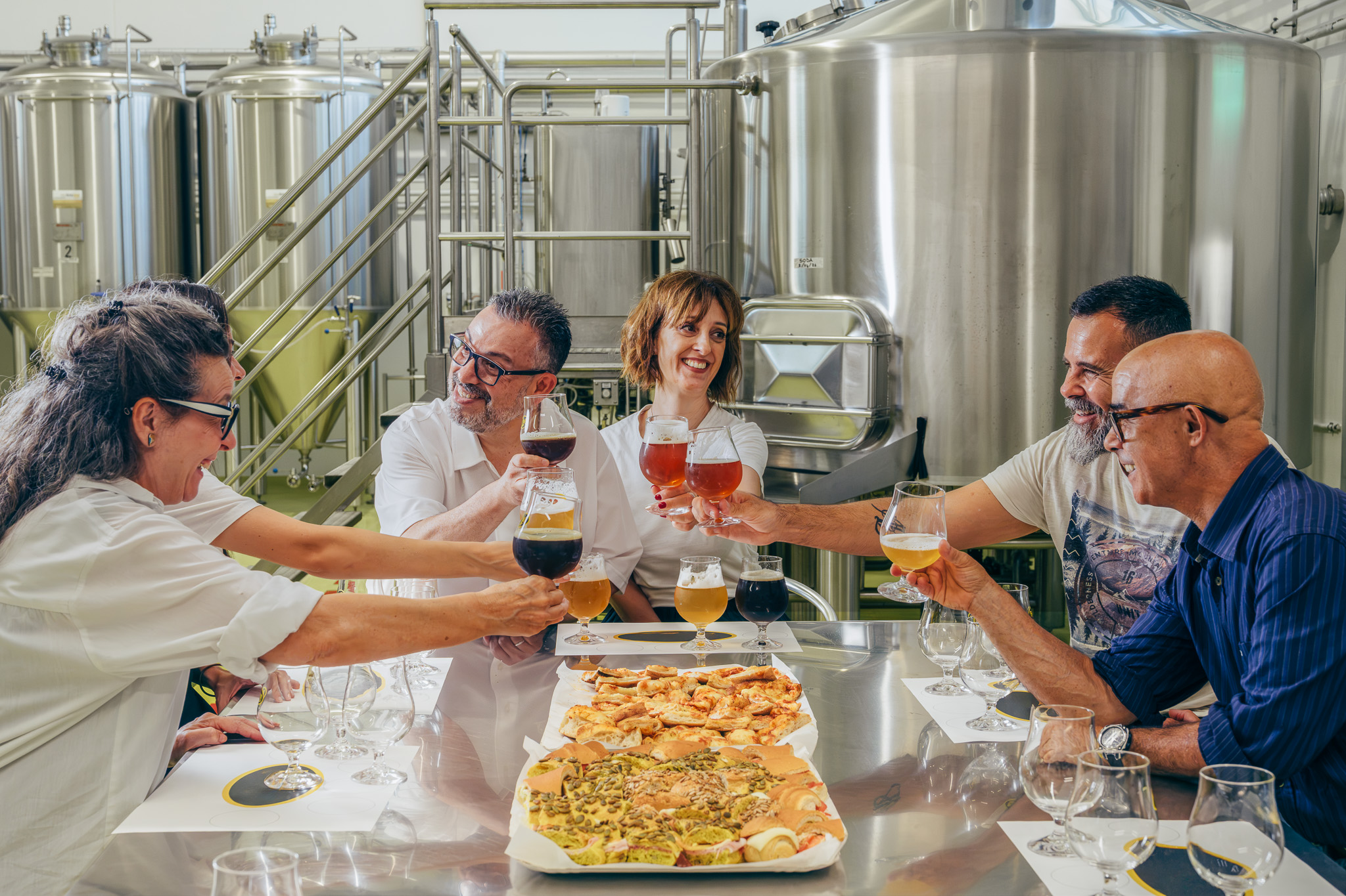 Un gruppo di persone brinda con bicchieri di birra davanti a un tavolo con cibo e birre in un ambiente industriale.