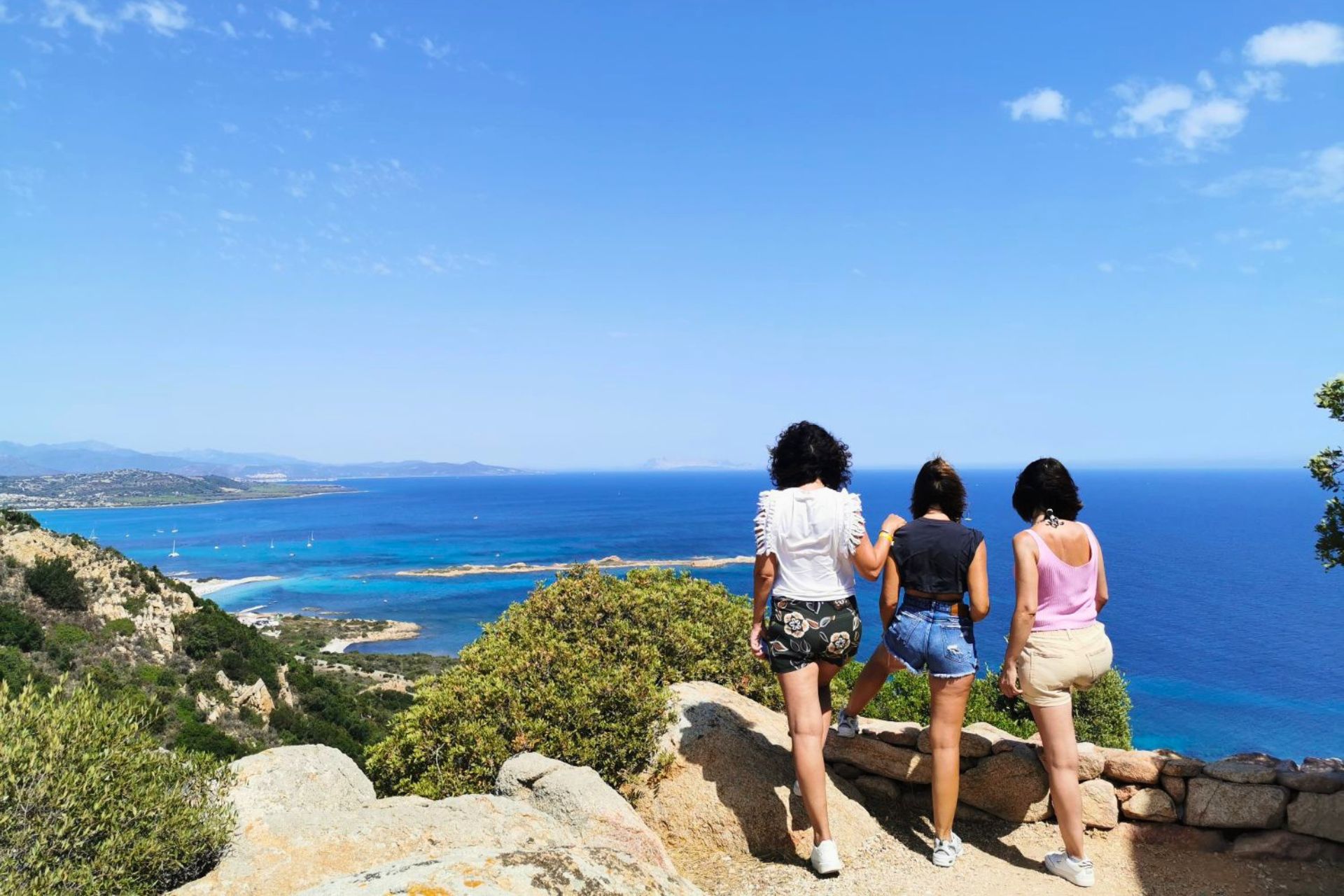 Trois femmes debout sur une falaise surplombant la mer bleue avec quelques îles et plages au loin