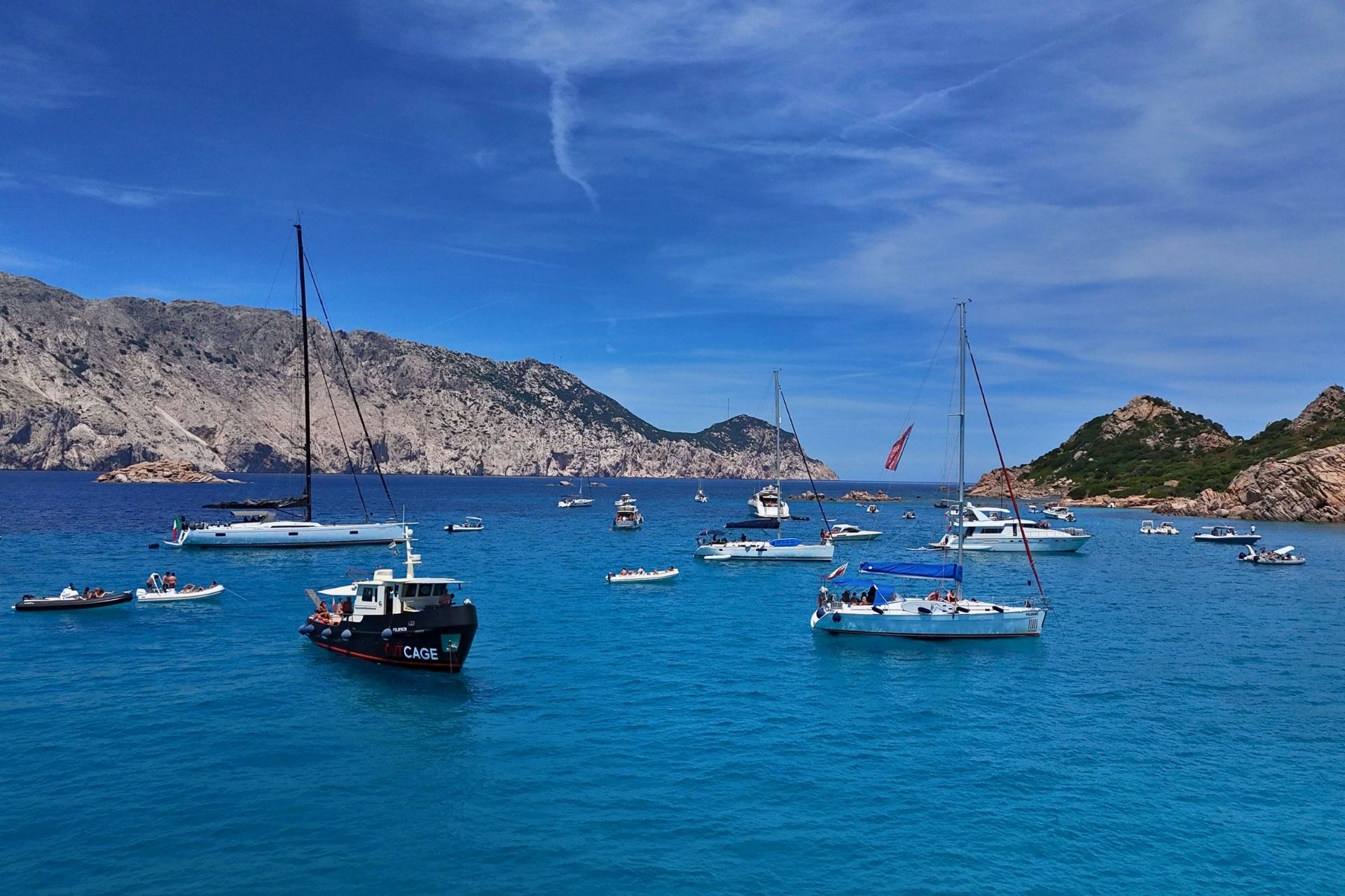 Imagen de un grupo de veleros y pequeñas embarcaciones en un mar cristalino con montañas de fondo.