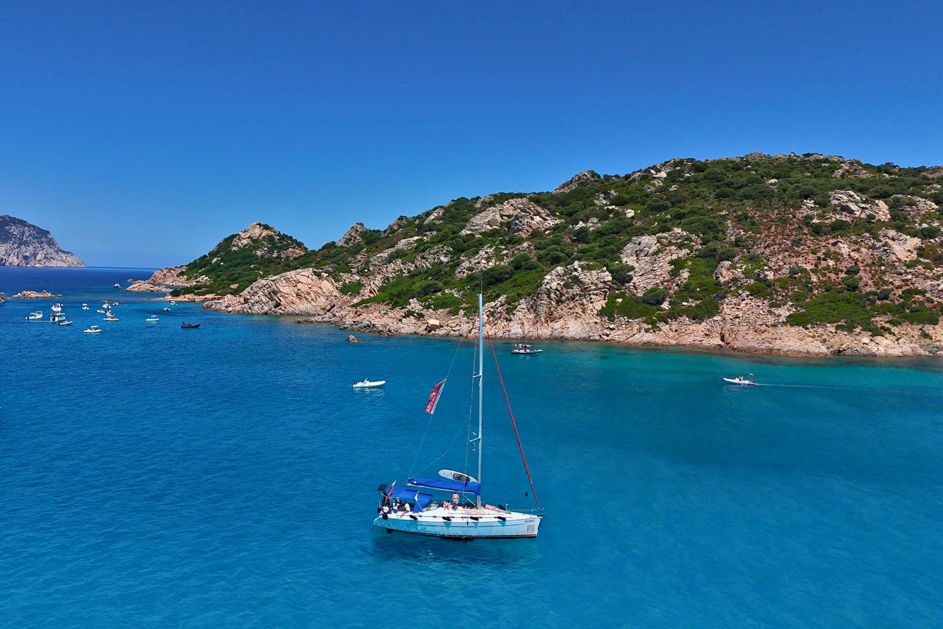 Un velero navega en aguas cristalinas y azules, con islotes y acantilados al fondo. Hay varios otros barcos a la vista, algunos con banderas izadas.