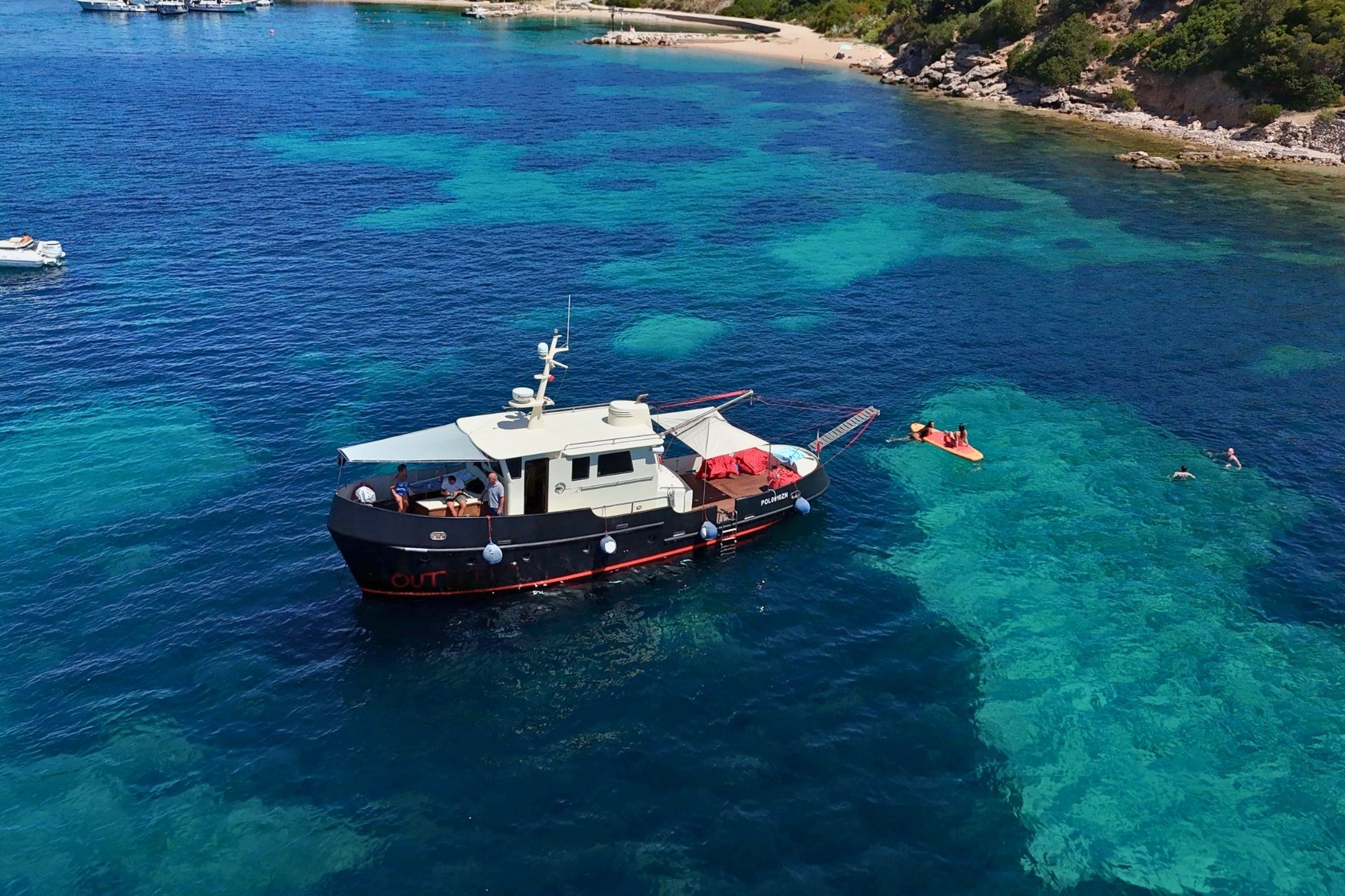 Un bateau d'époque navigue dans une eau claire avec des personnes faisant de la plongée en apnée et d'autres nageant dans une eau cristalline.