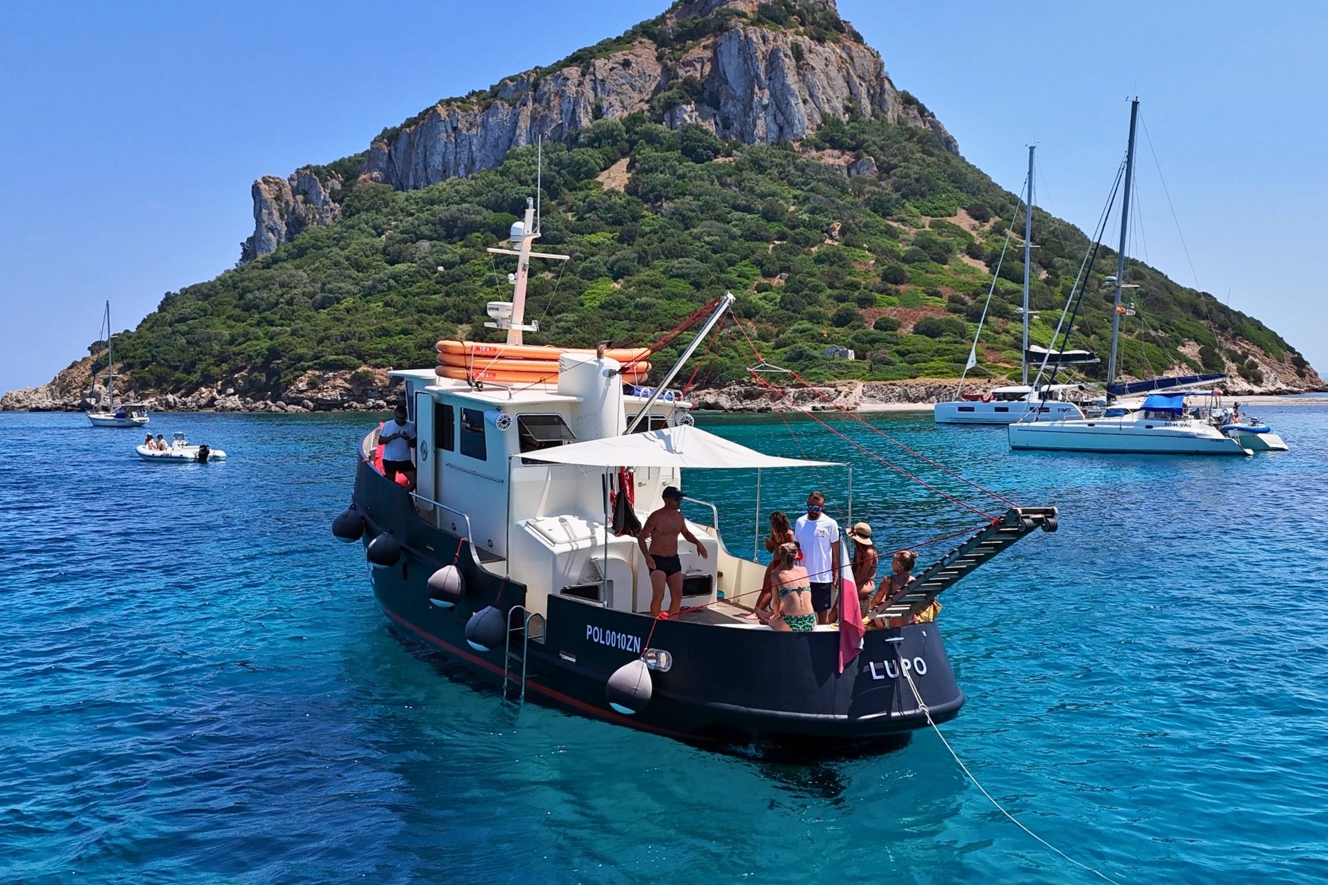 Un bateau d'époque navigue dans des eaux cristallines avec des personnes à bord, sur une côte verdoyante avec des falaises en arrière-plan.