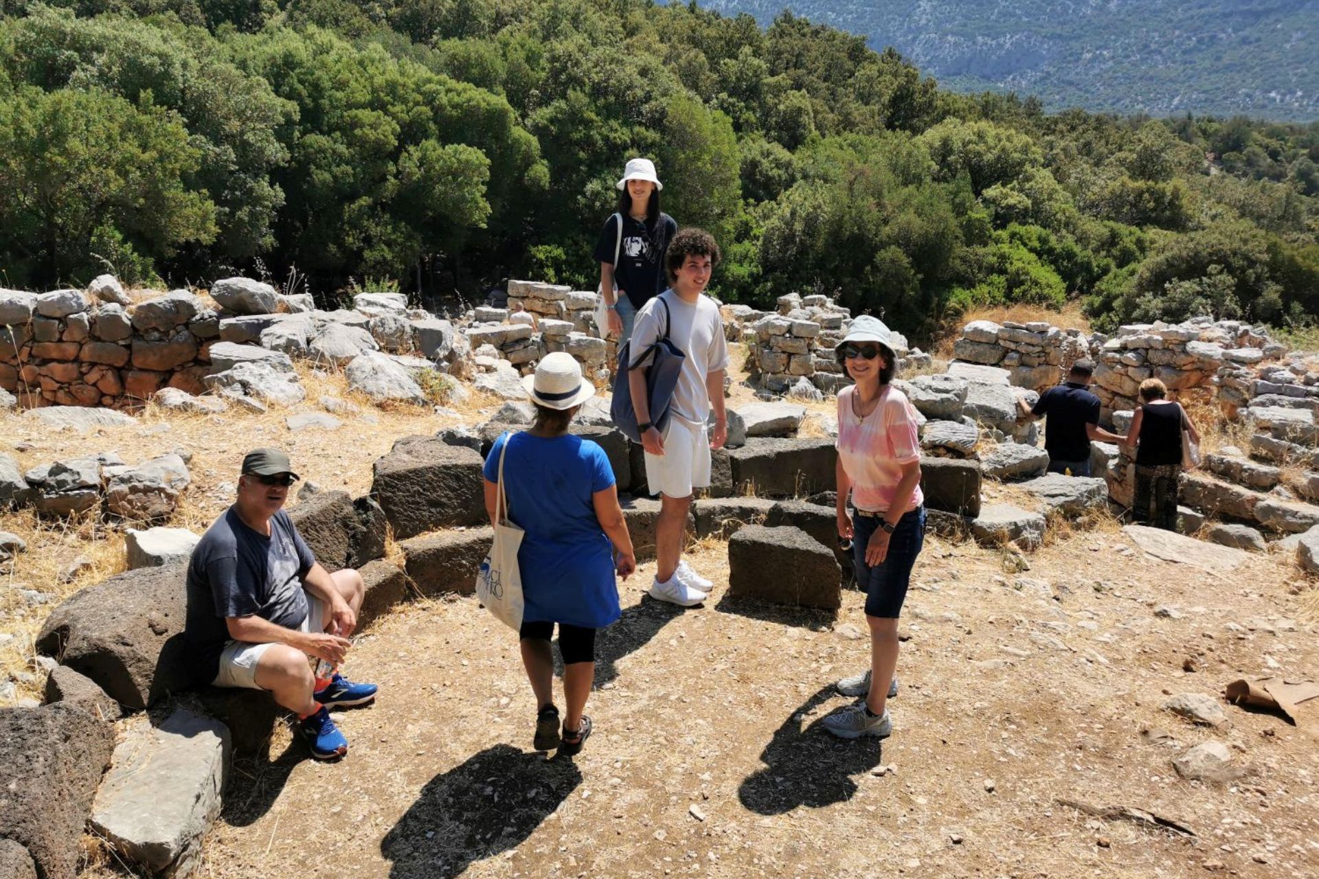 A group of people visit an open-air archaeological site with stone ruins and green vegetation around it.
