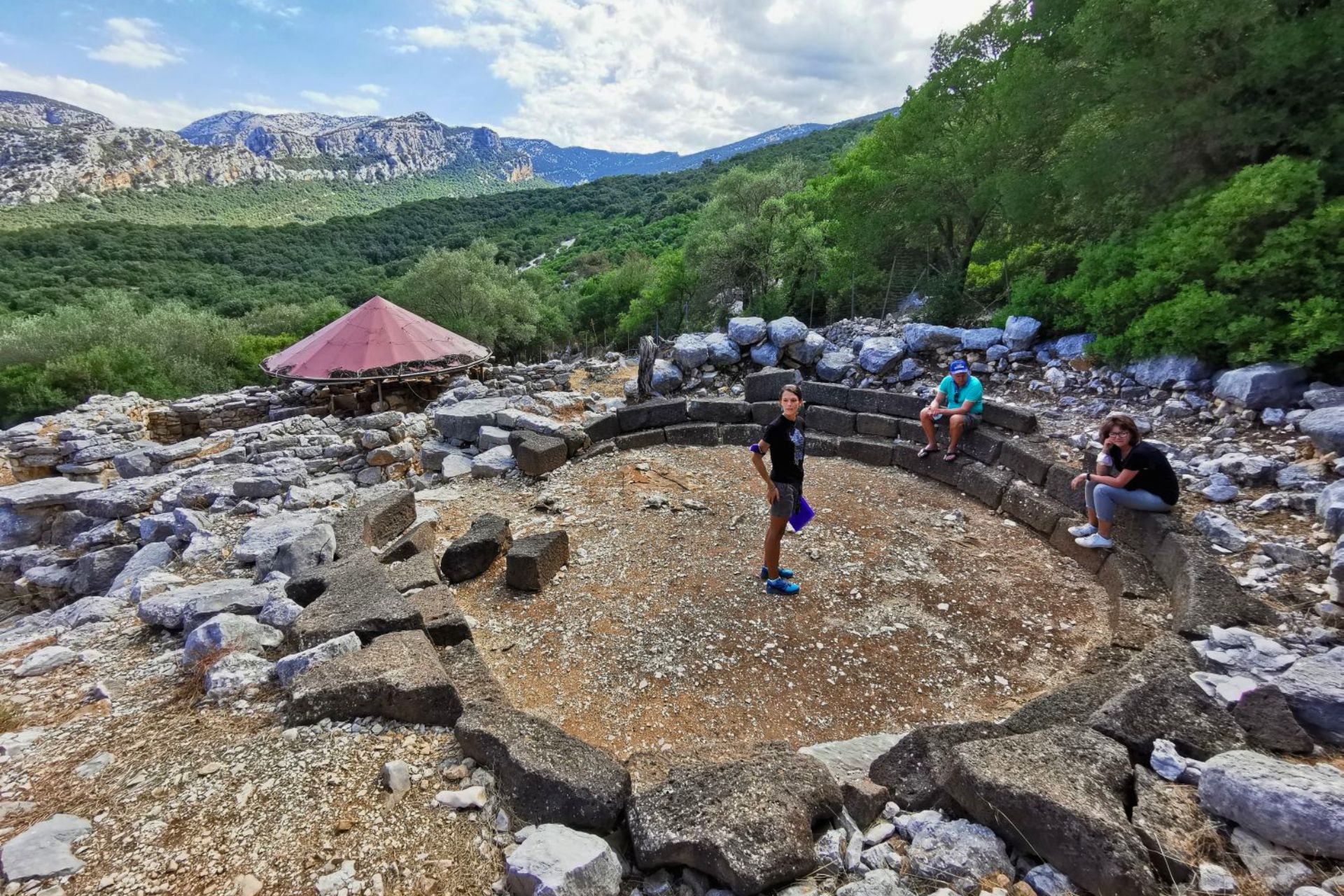 Image of a group of people at an outdoor archaeological site, with stone ruins and a red shelter