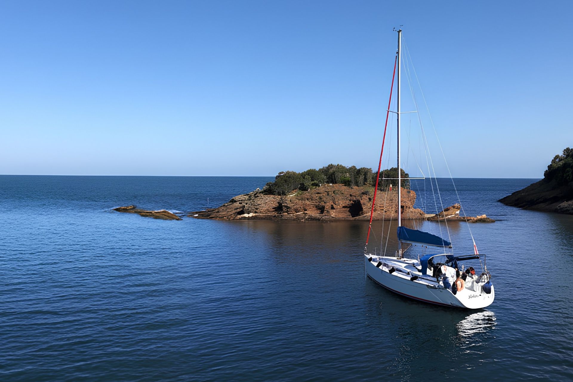 Un velero navega por un mar azul claro, con una isla rocosa al fondo y algunas rocas sumergidas visibles en el agua.
