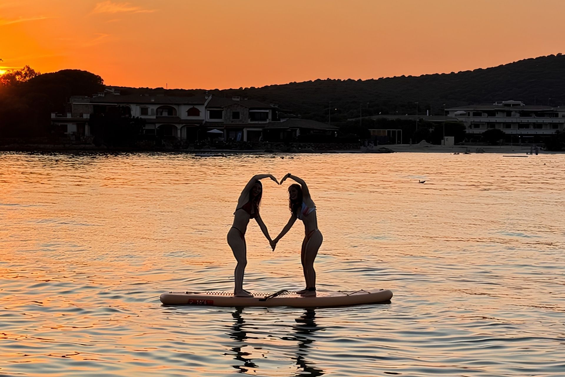 Dos mujeres en bikini sobre una tabla de surf formando un corazón con las manos sobre el fondo de una puesta de sol.