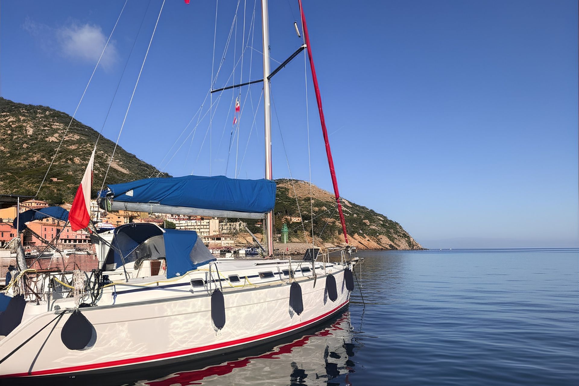 Un velero blanco amarrado en un puerto con montañas de fondo y mar en calma.