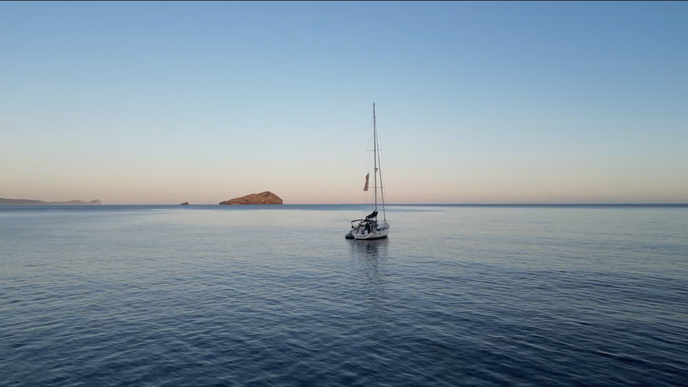 Una barca a vela naviga in mare aperto con il sole che cala all'orizzonte. Sullo sfondo si intravedono alcune isole.