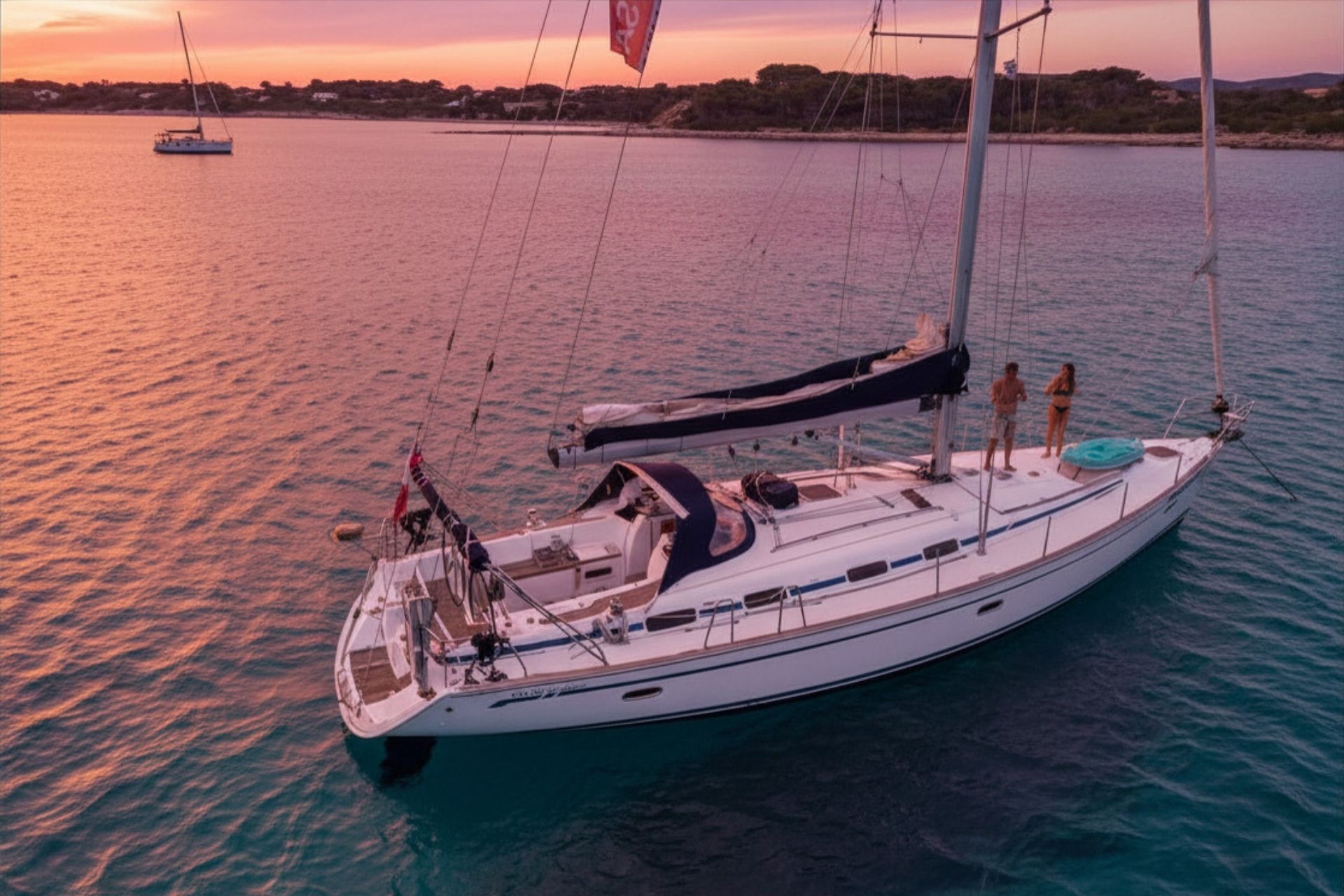 Barca a vela con passeggeri sul mare al tramonto, sullo sfondo un'altra barca e un paesaggio costiero.