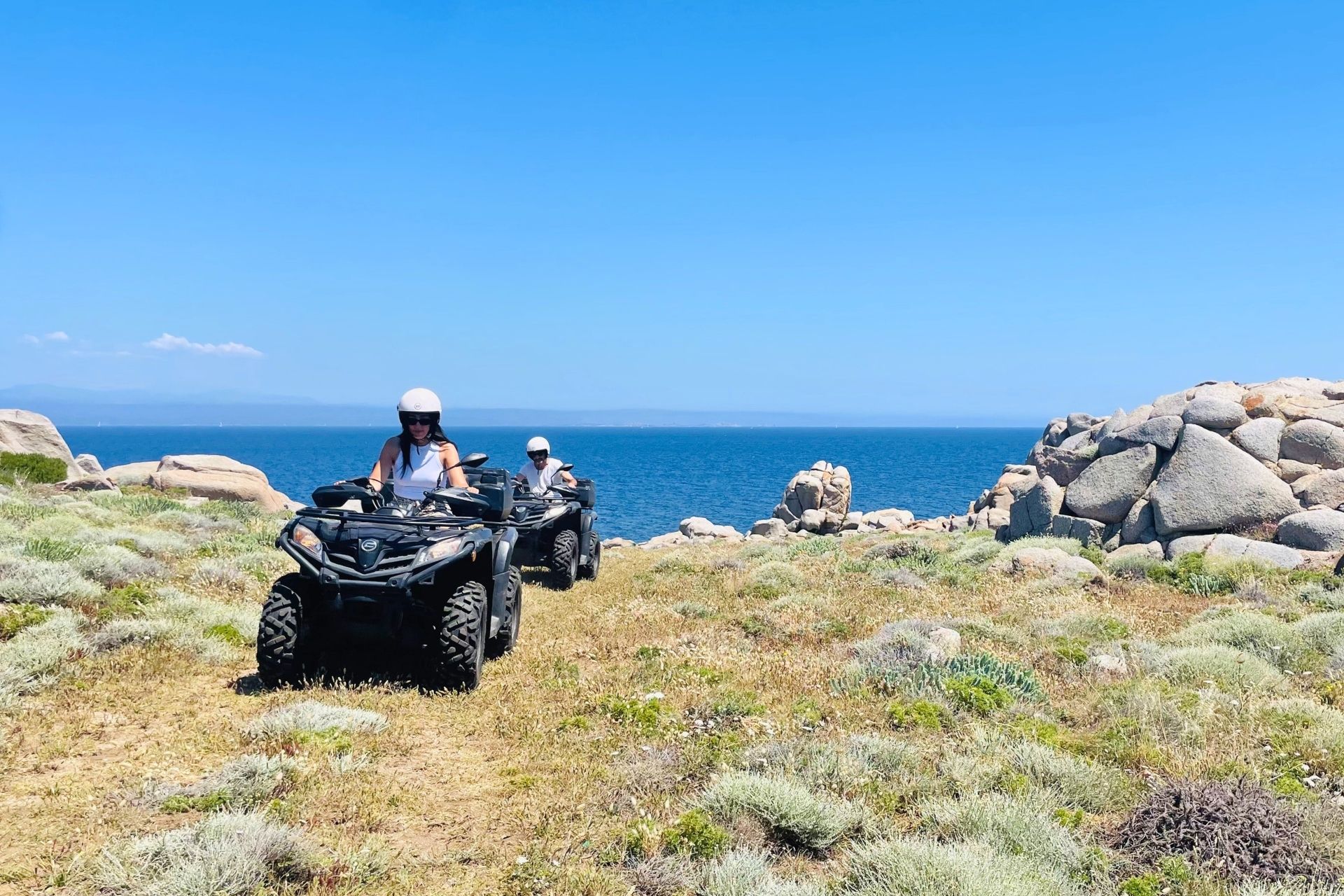 Due persone su un quad su un sentiero di campagna vicino al mare, con rocce e vegetazione tipica del luogo.