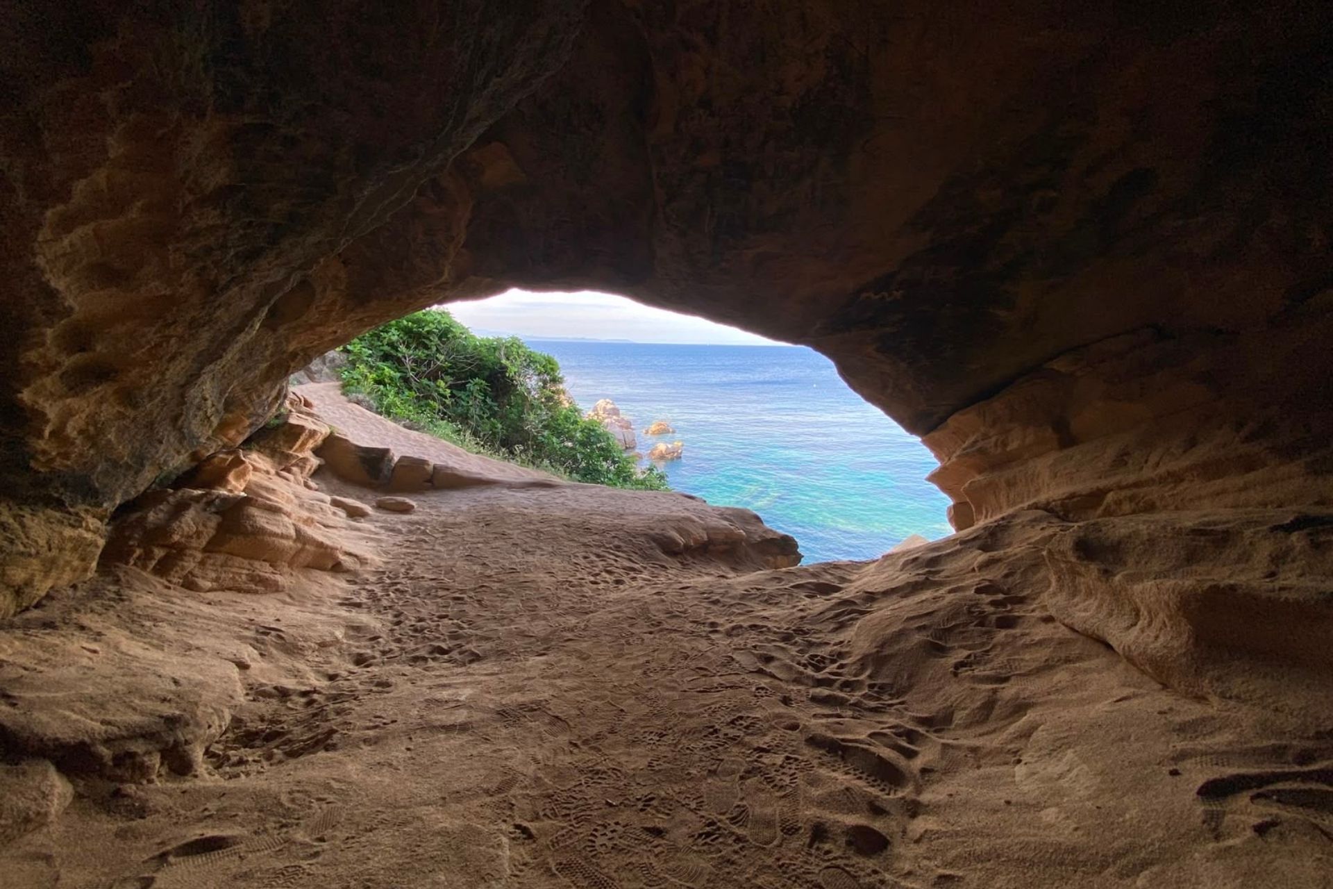 Una grotta con vista sul mare e sulla costa, con tracce di passi sulla sabbia e roccia.