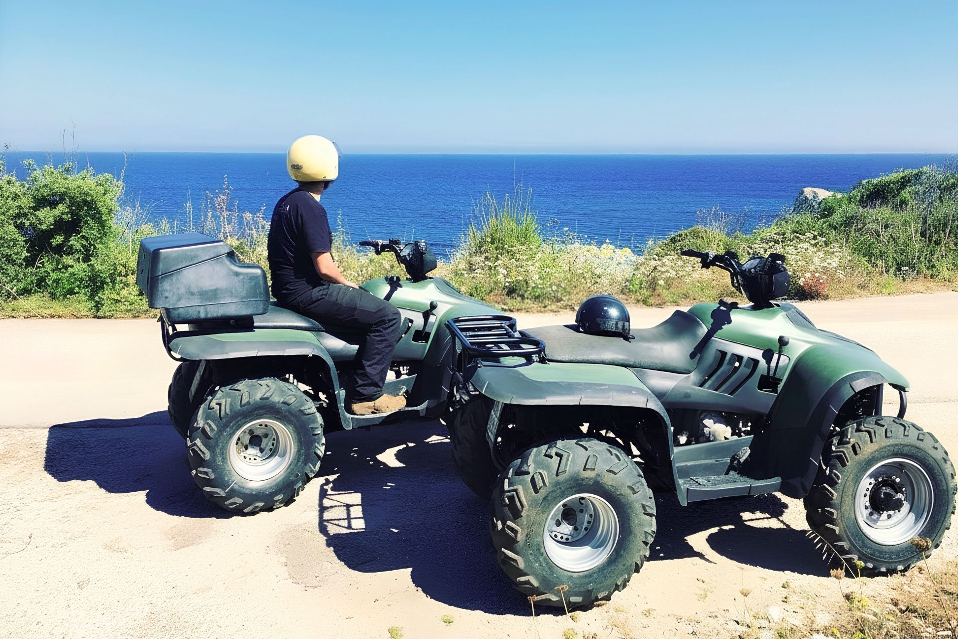Due persone in quad, con caschi, in una strada panoramica con vista sul mare e sulla natura circostante.