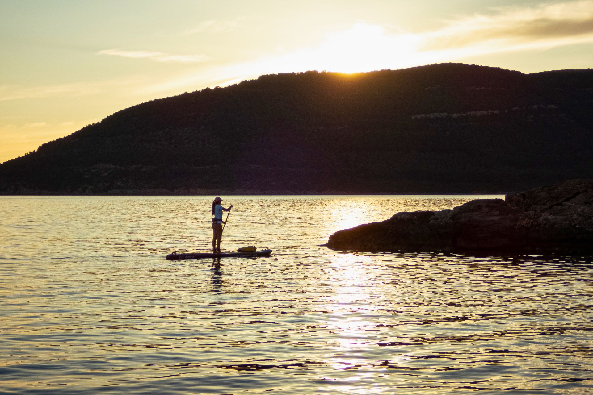 Une personne debout sur une planche de SUP dans l'eau calme avec une montagne en arrière-plan au coucher du soleil