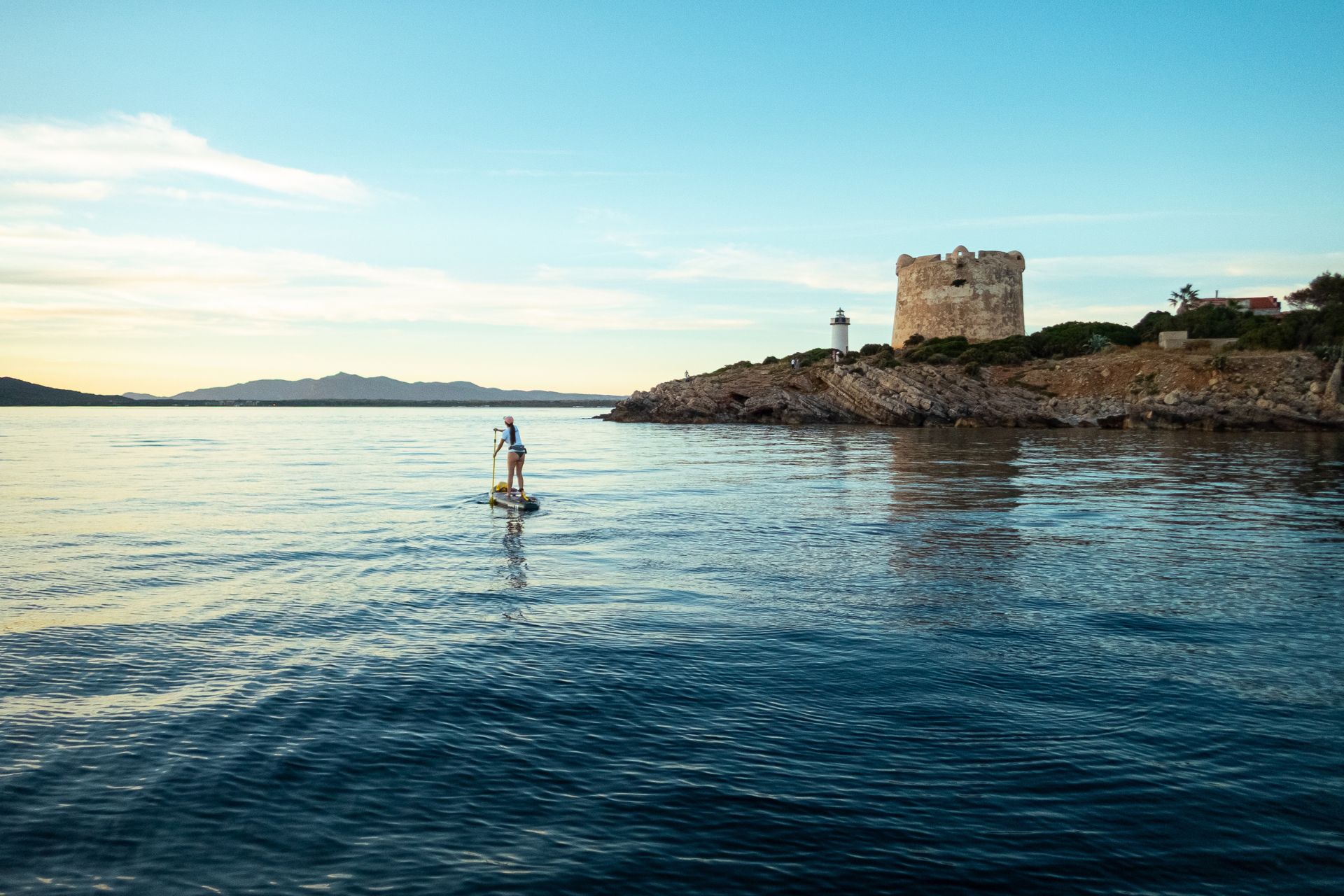 Une personne debout sur une planche de SUP en mer, en arrière-plan une île avec un phare et un château