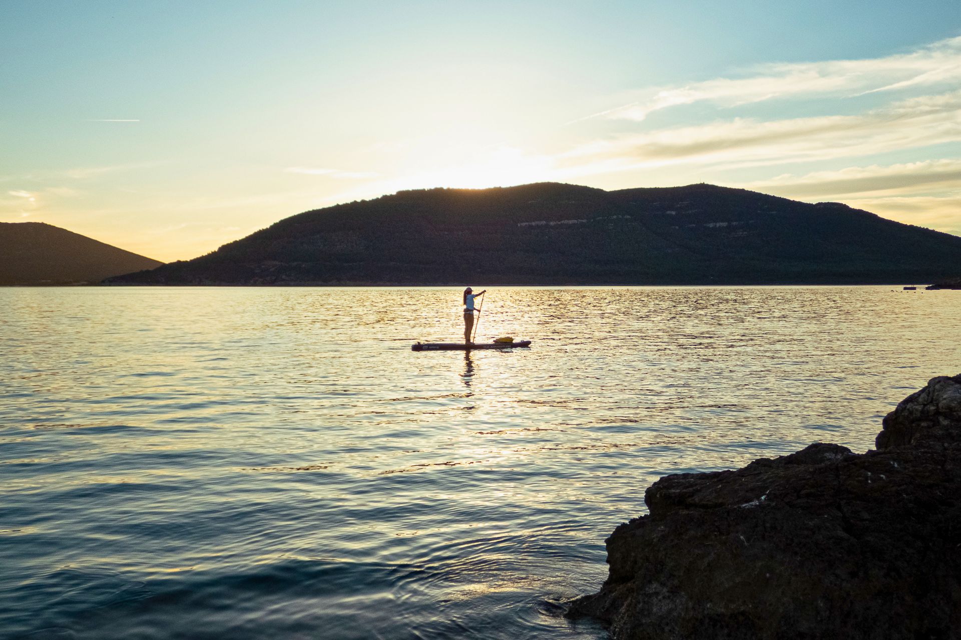 Une personne debout sur une planche de SUP en eau calme avec des montagnes en arrière-plan au coucher du soleil