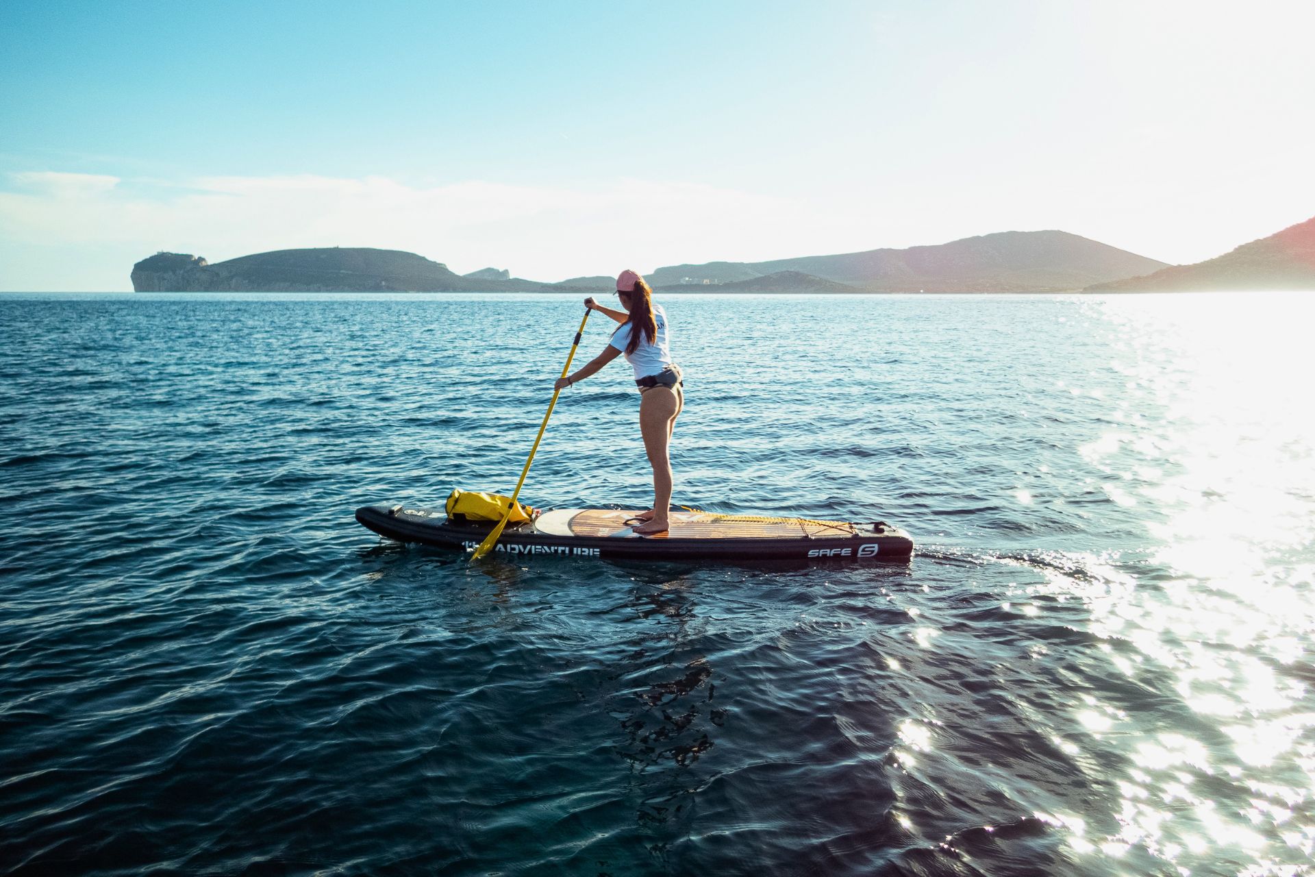 Une femme debout sur une planche de SUP dans l'océan au coucher du soleil, avec une île en arrière-plan et des montagnes au loin. La planche de SUP a une inscription blanche et jaune