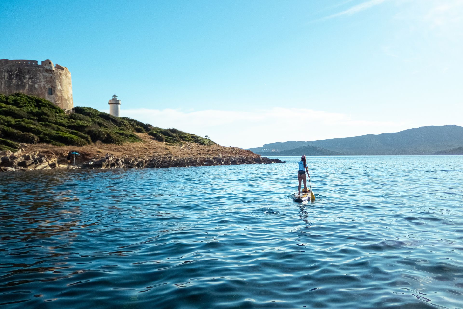 Une personne debout sur une planche de SUP dans l'eau, avec une ancienne tour en arrière-plan et un paysage naturel autour