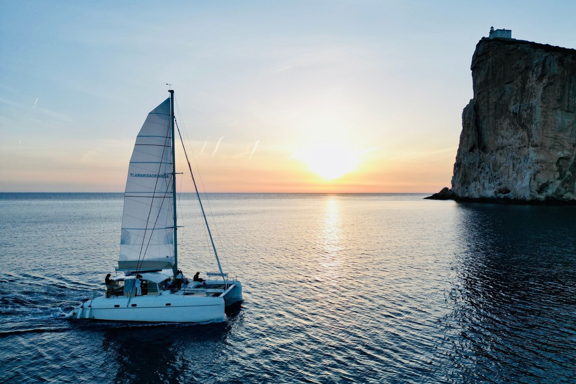 Navegación en catamarán al atardecer con gente a bordo y una roca al fondo