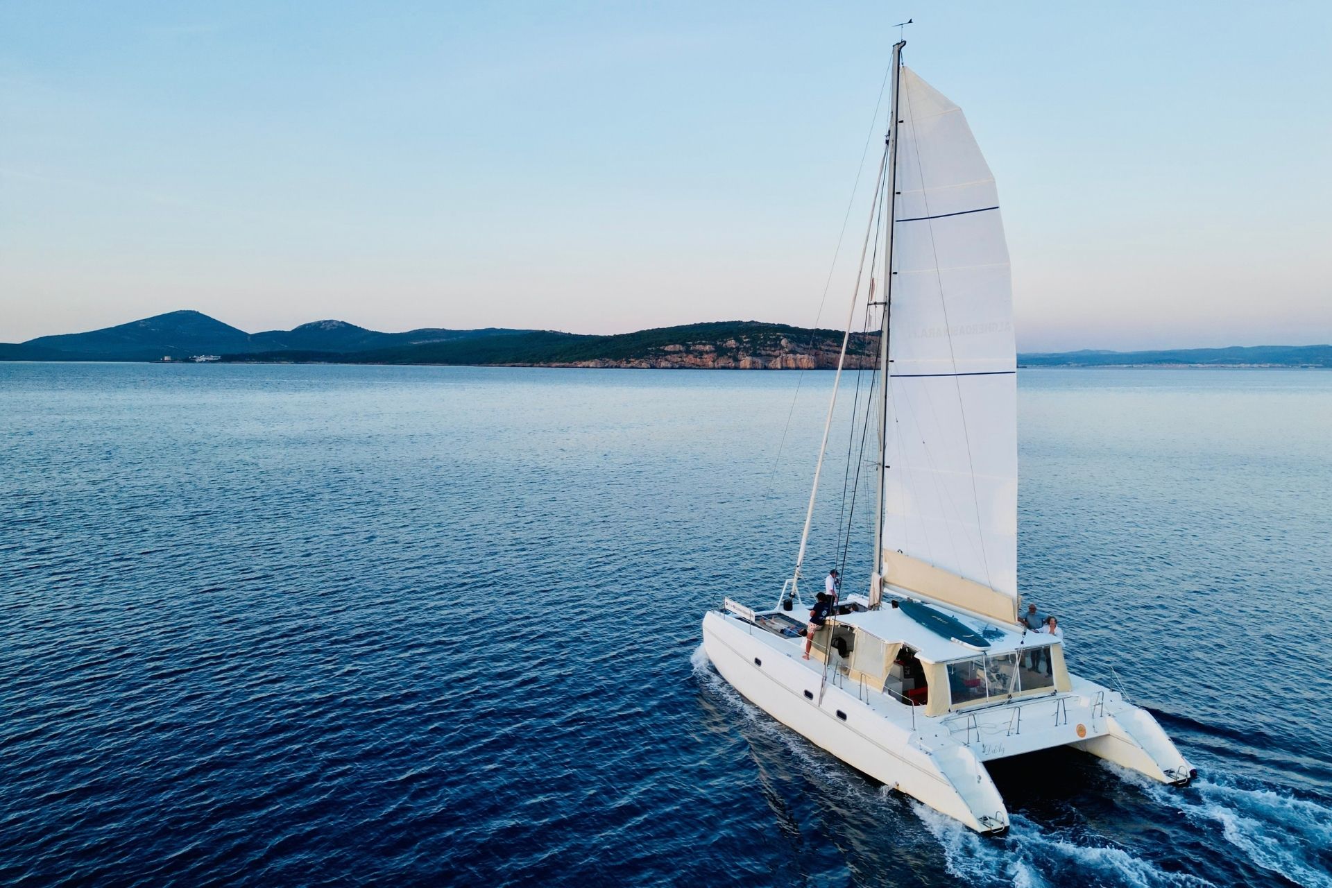 Un catamarán navega por la costa de Alghero durante la puesta de sol, con un grupo de personas a bordo.