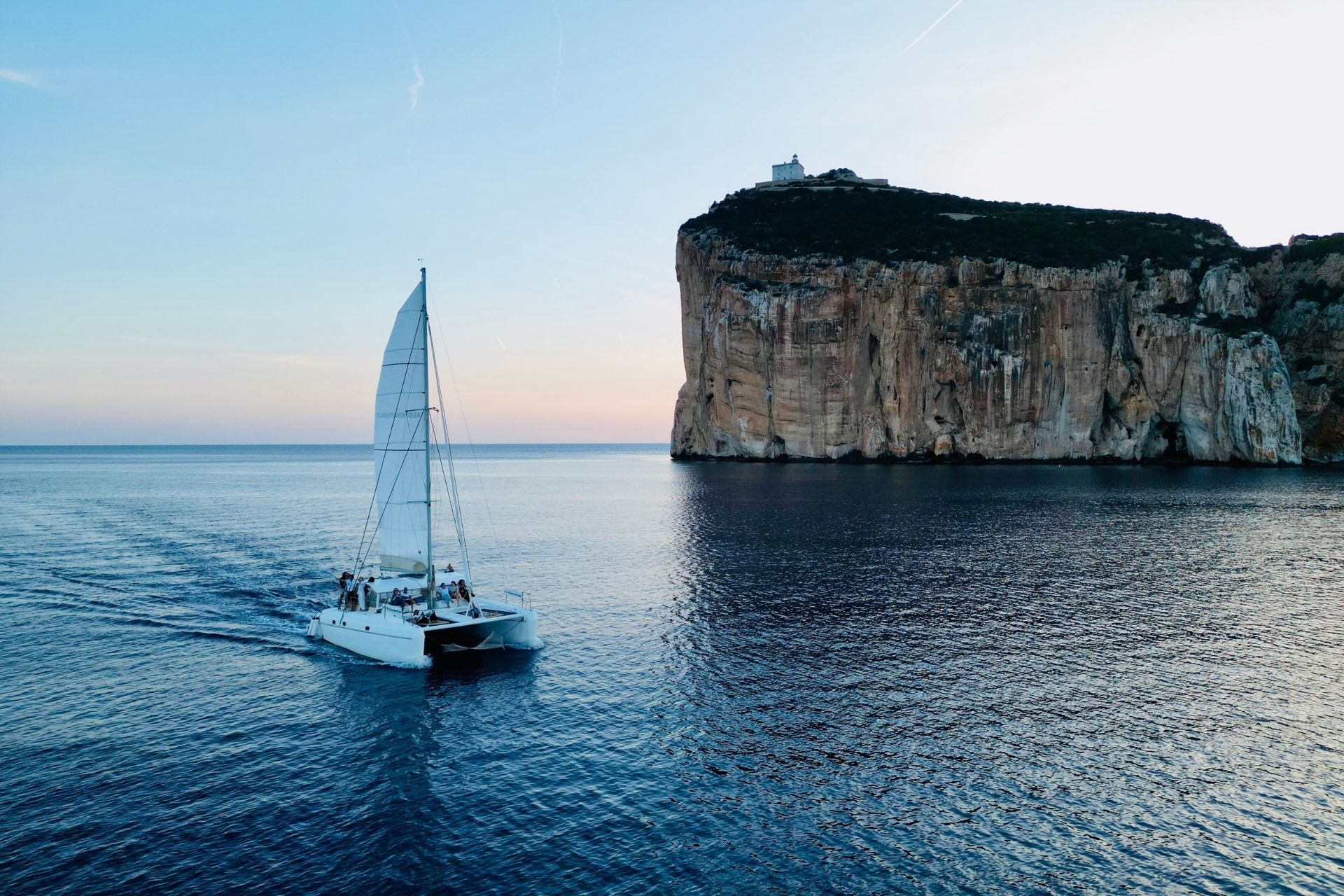 Un catamarán navega cerca de un acantilado con un faro en la parte superior, durante la puesta de sol.