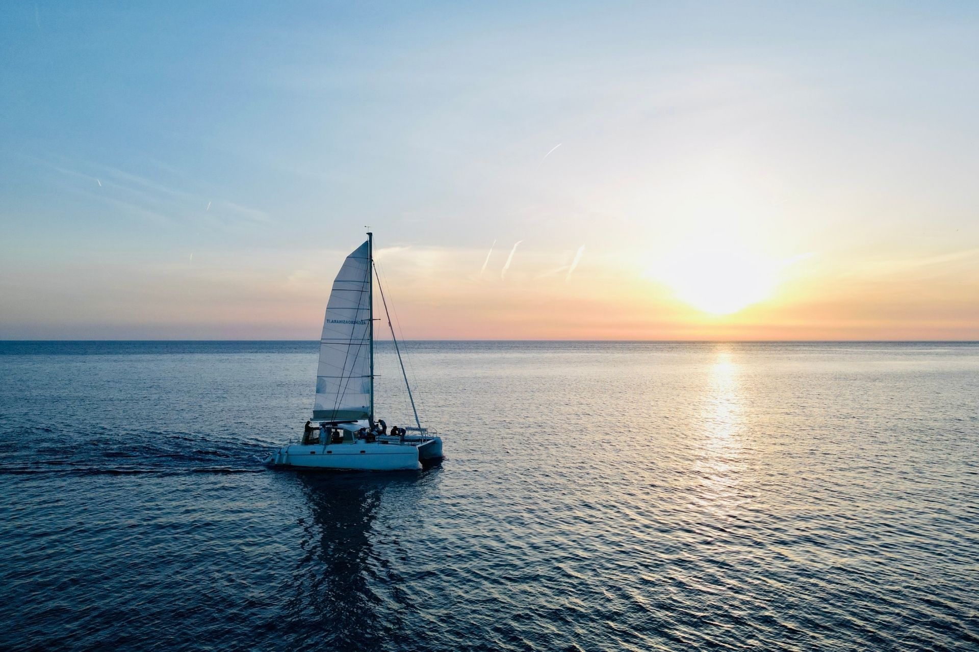 Imagen de un catamarán navegando por el mar al anochecer con un grupo de personas a bordo. La puesta de sol se refleja en las aguas tranquilas. El paisaje circundante muestra una costa con acantilados y vegetación. El ambiente evoca tranquilidad y belleza natural.