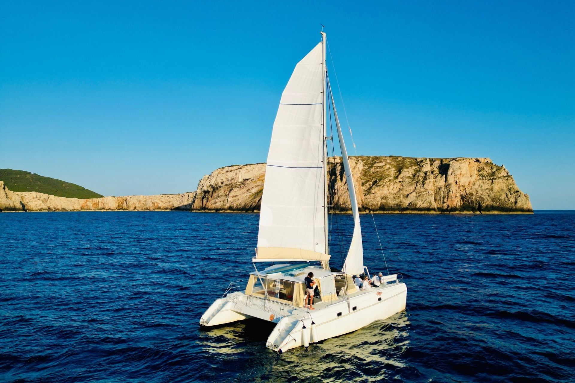Un catamarán navega en mar abierto con una isla rocosa al fondo, bajo un cielo azul despejado.
