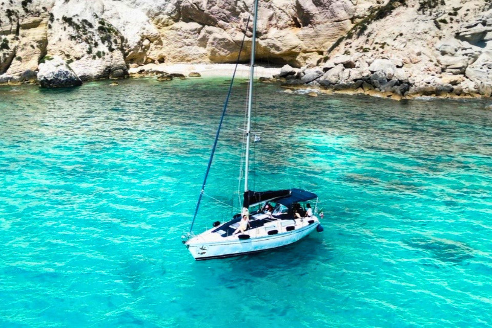 Image of a sailboat moored in crystal clear waters with people on board and rocks in the background