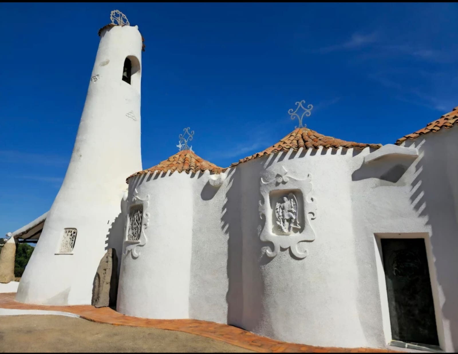Image of a white building with red roof and artistic decorations, located in a natural landscape with blue sky and clouds