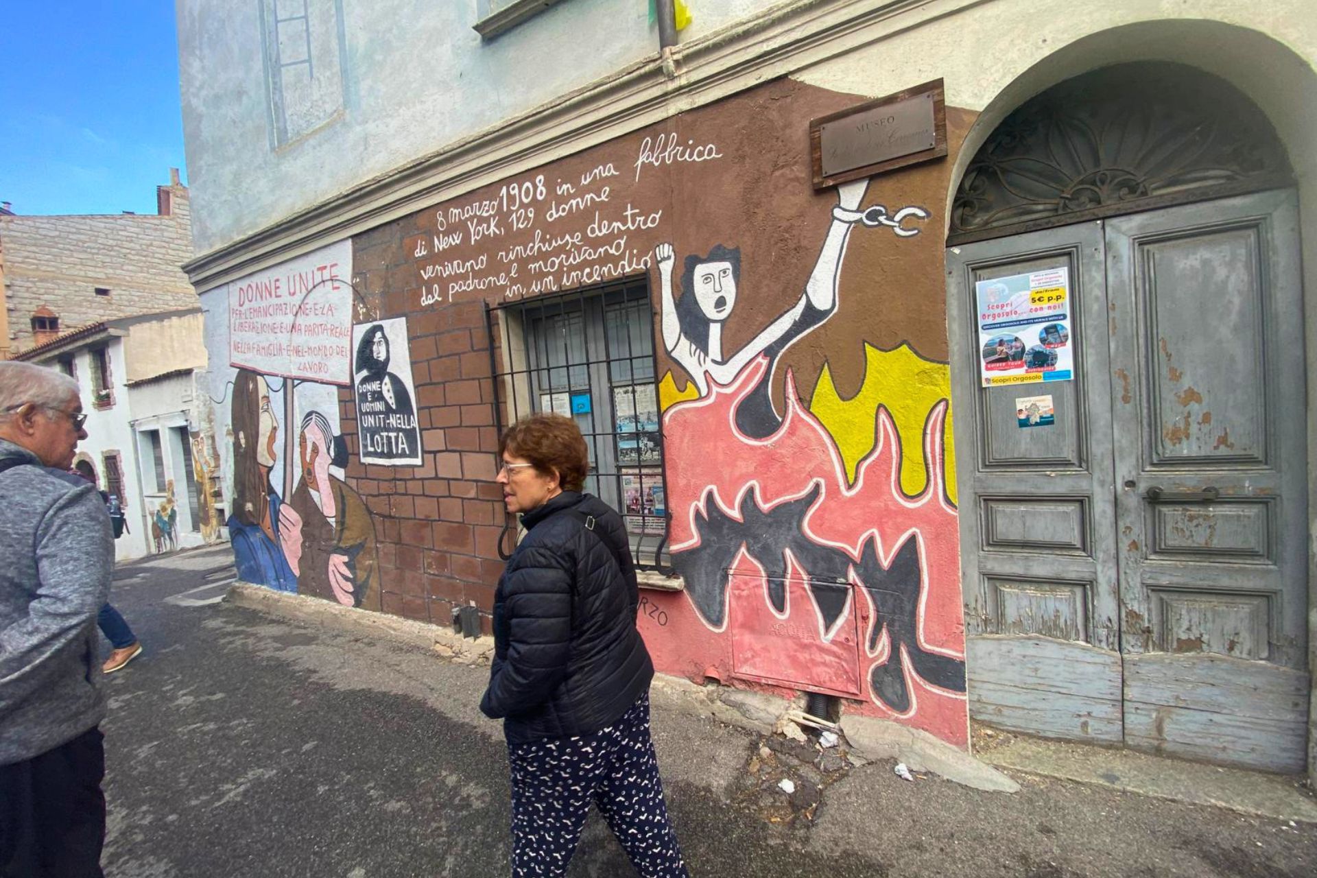Une femme marche devant un bâtiment orné de peintures murales représentant des personnages historiques et culturels de la Sardaigne