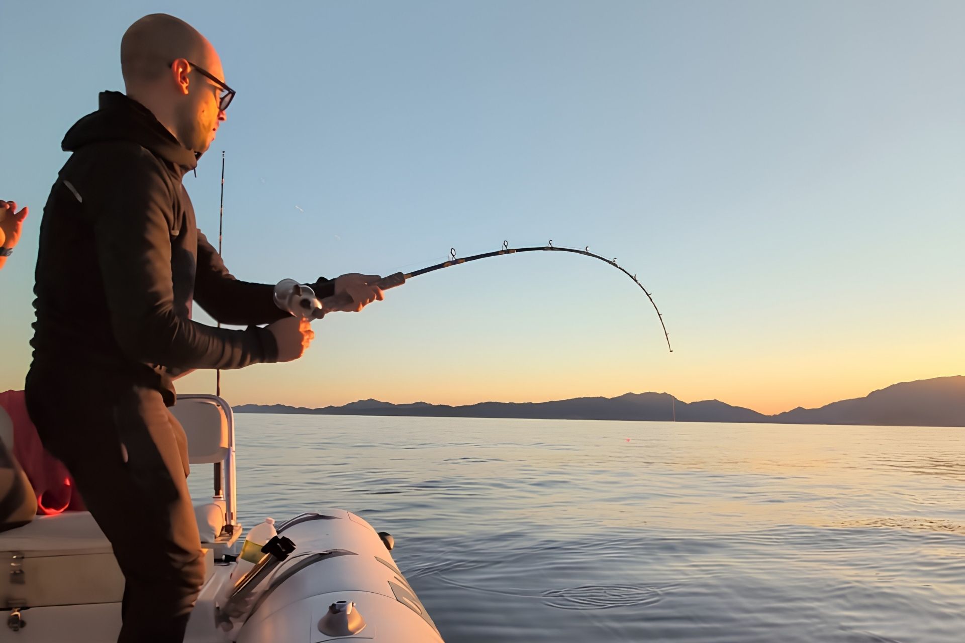 A man fishes in the sea at dusk from an equipped dinghy, wearing sportswear and sunglasses. In the background you can see mountains and a setting sky.