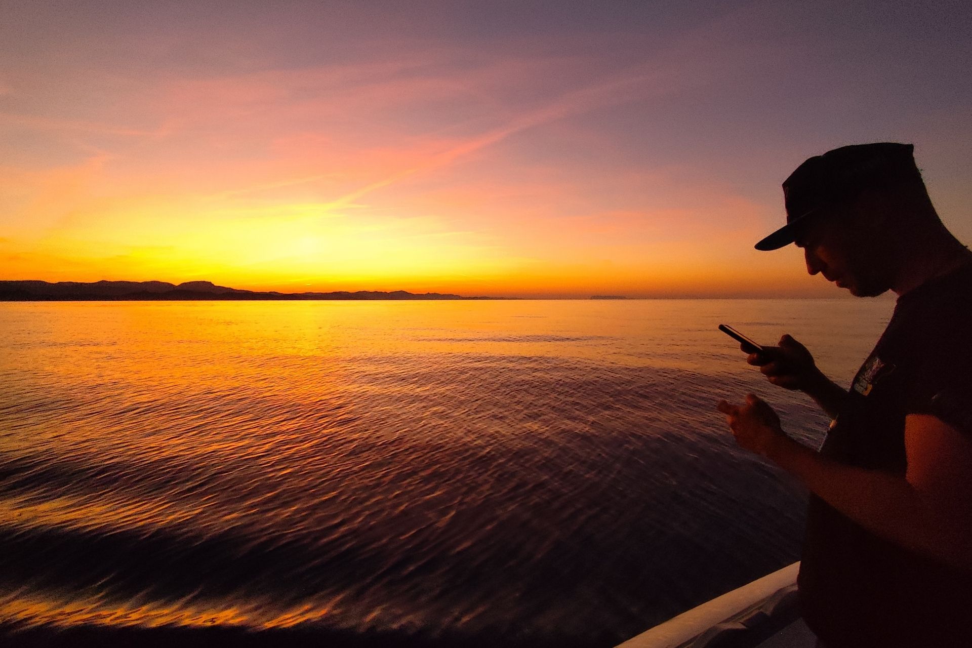 A man looks at his cell phone while fishing on a sailboat with a sunset in the background