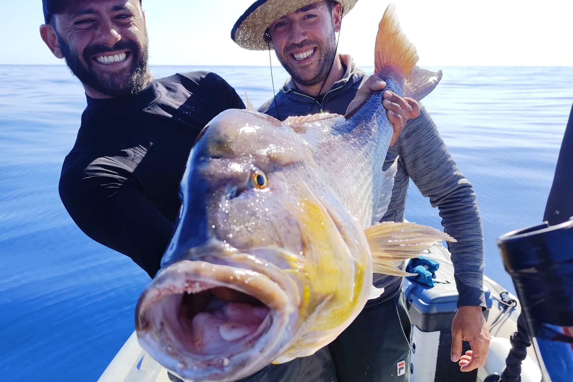 Two fishermen smile while holding a large fish in their hands.