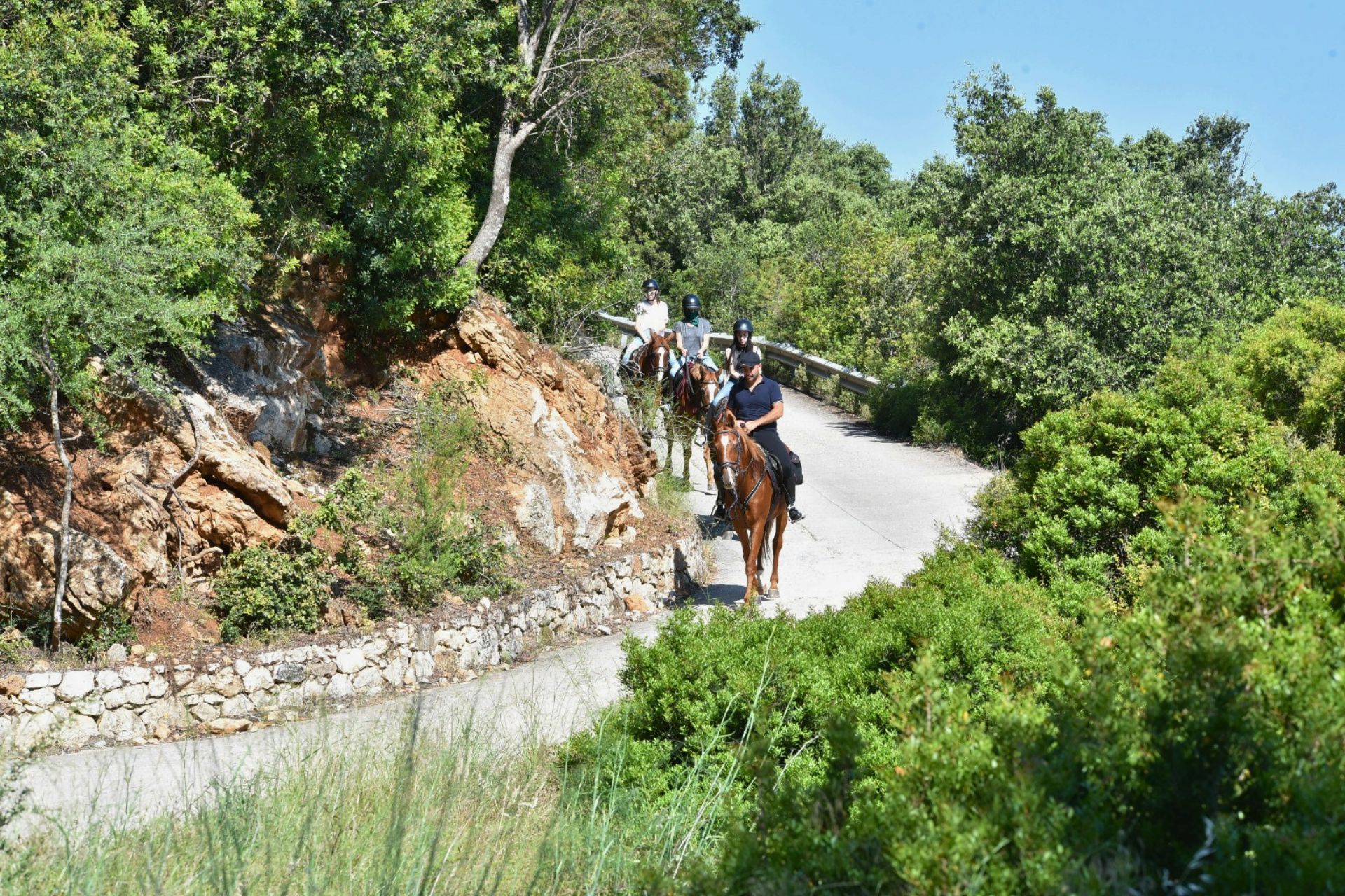 Persone a cavallo in una valle con vegetazione rigogliosa e rocce, sotto un cielo azzurro