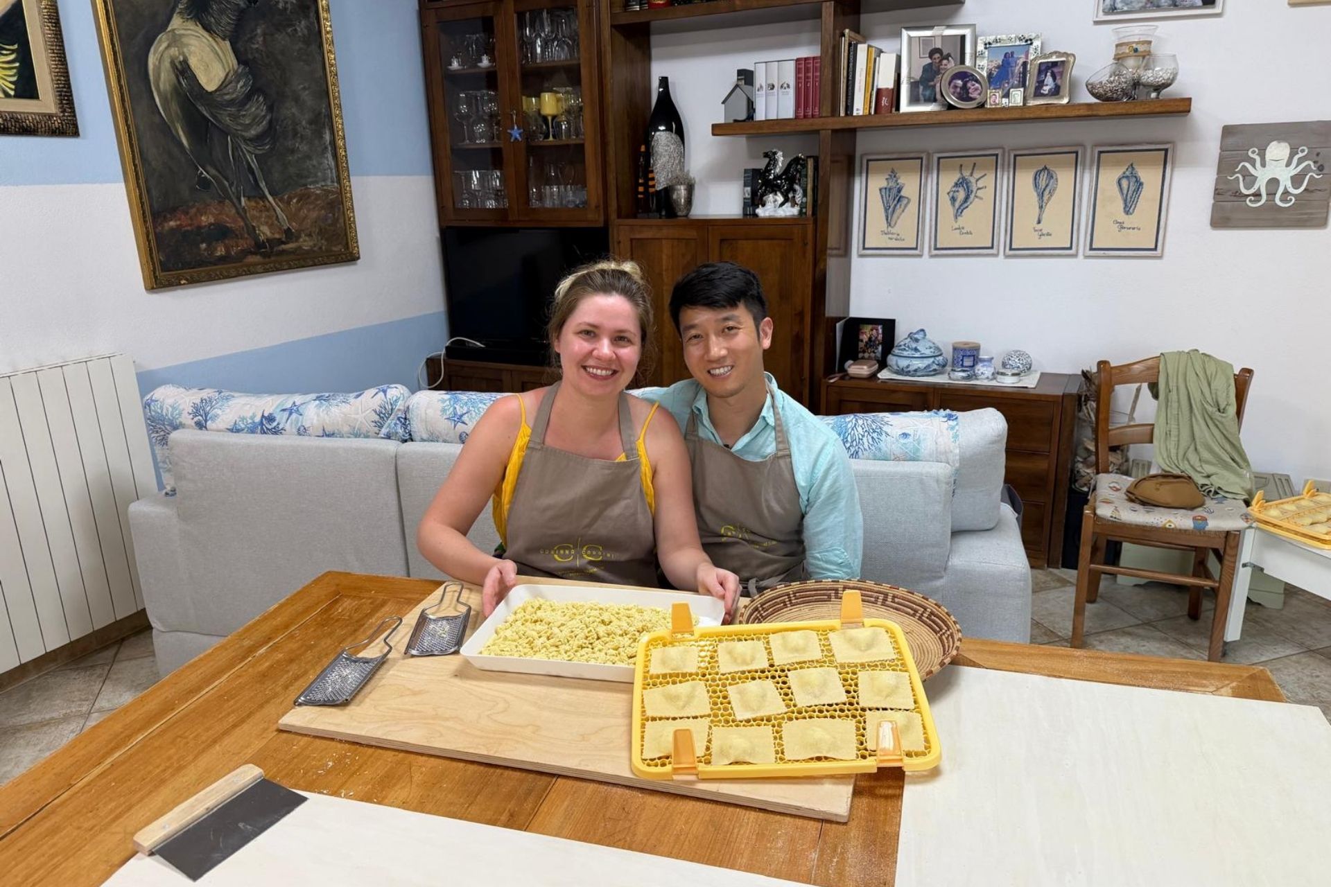 Two people in a living room, with a table and a plate of fresh pasta, smile. In the background, a sofa, a shelf with objects and paintings and an open window. A fresh Sardinian pasta workshop in Arzachena.