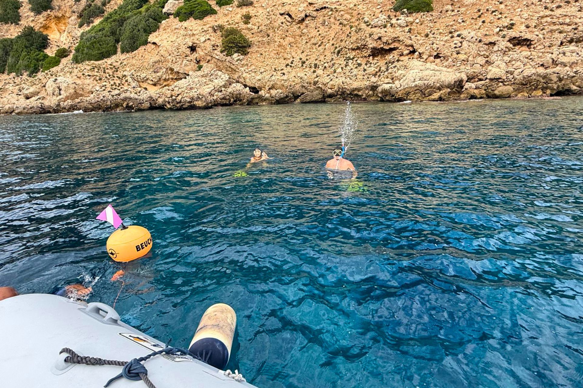 Tre persone fanno snorkeling in mare, con un gommone in agguato a sinistra. Sullo sfondo si intravede una costa rocciosa e verdeggiante. Un'esperienza di snorkeling organizzata ad Olbia