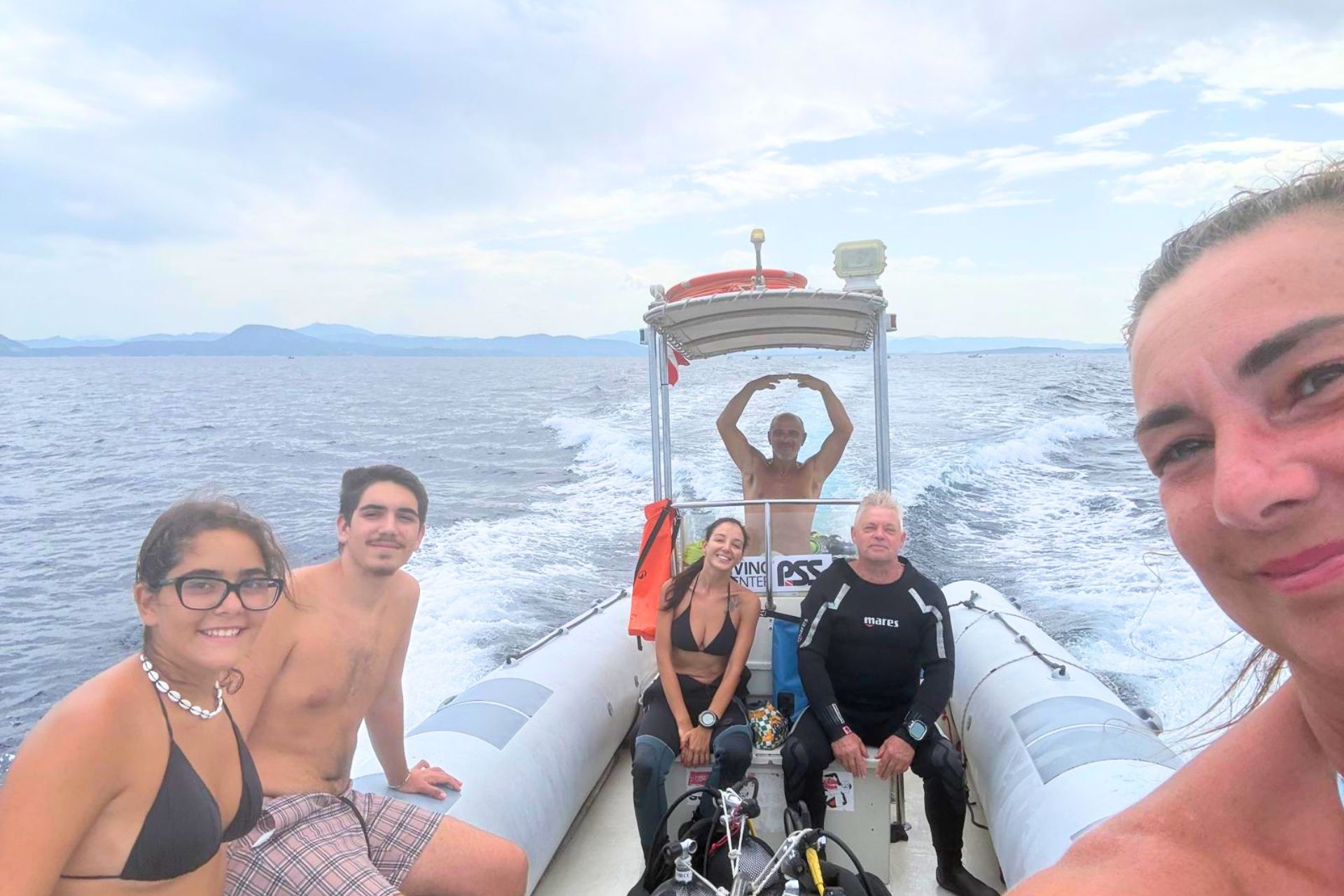 Gruppo di persone su un gommone in mare, con un uomo in piedi sul bordo e una donna che sorride alla telecamera