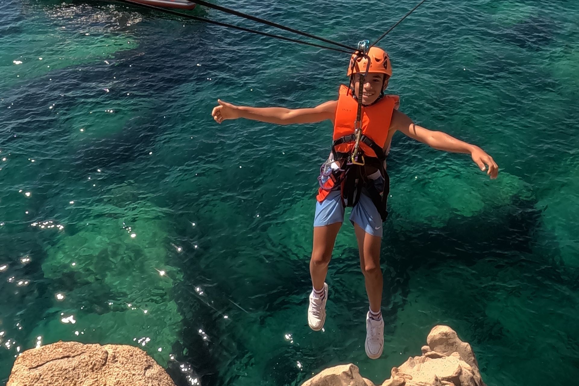 Un giovane in tuta e casco si lancia da un'altura verso il mare azzurro e cristallino, con un'espressione di gioia e sorpresa sul viso.