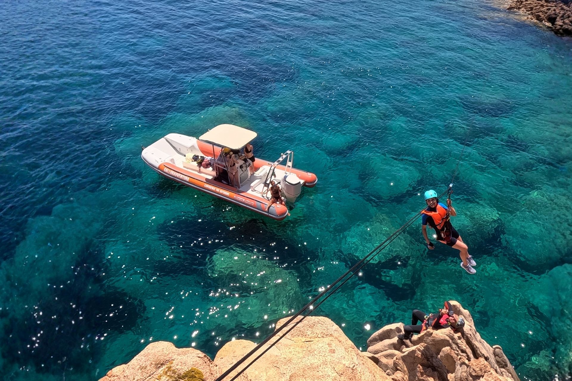 Una persona in tuta da coasteering si lancia in acqua da una scogliera, con un'imbarcazione in attesa a poca distanza.