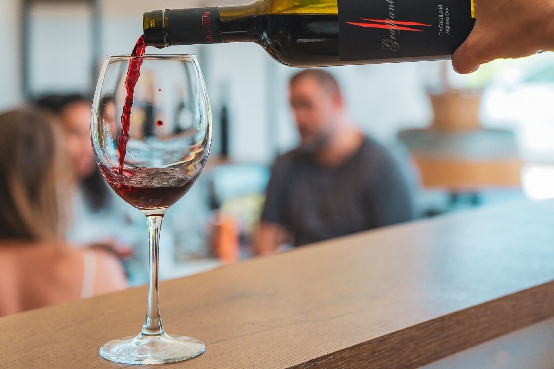 Une personne verse du vin rouge dans un verre dans une pièce avec d'autres clients assis à des tables.