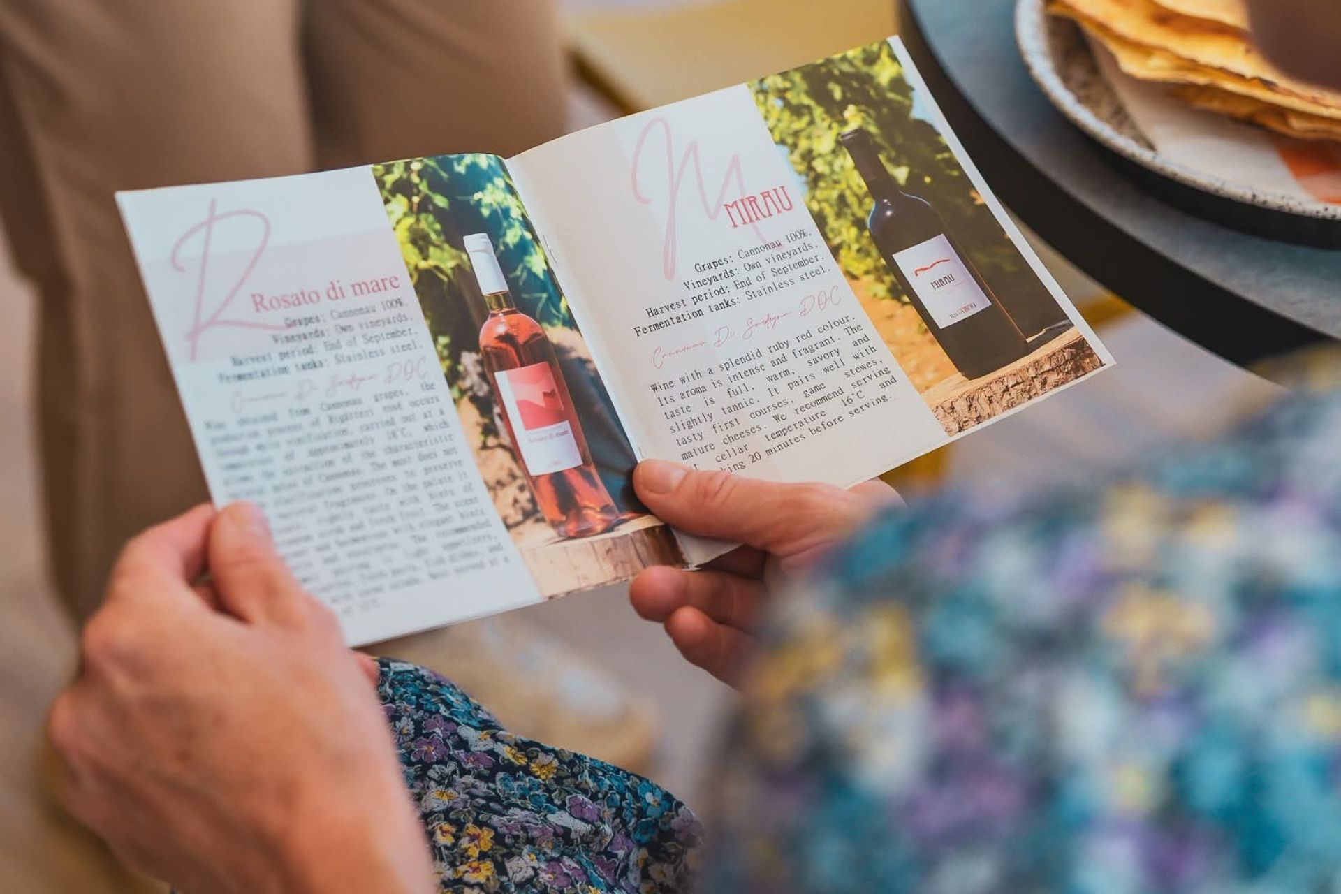 Personne lisant un menu de vins avec des images de bouteilles et de vignobles. Au fond, une table avec de la nourriture et un autre vin.