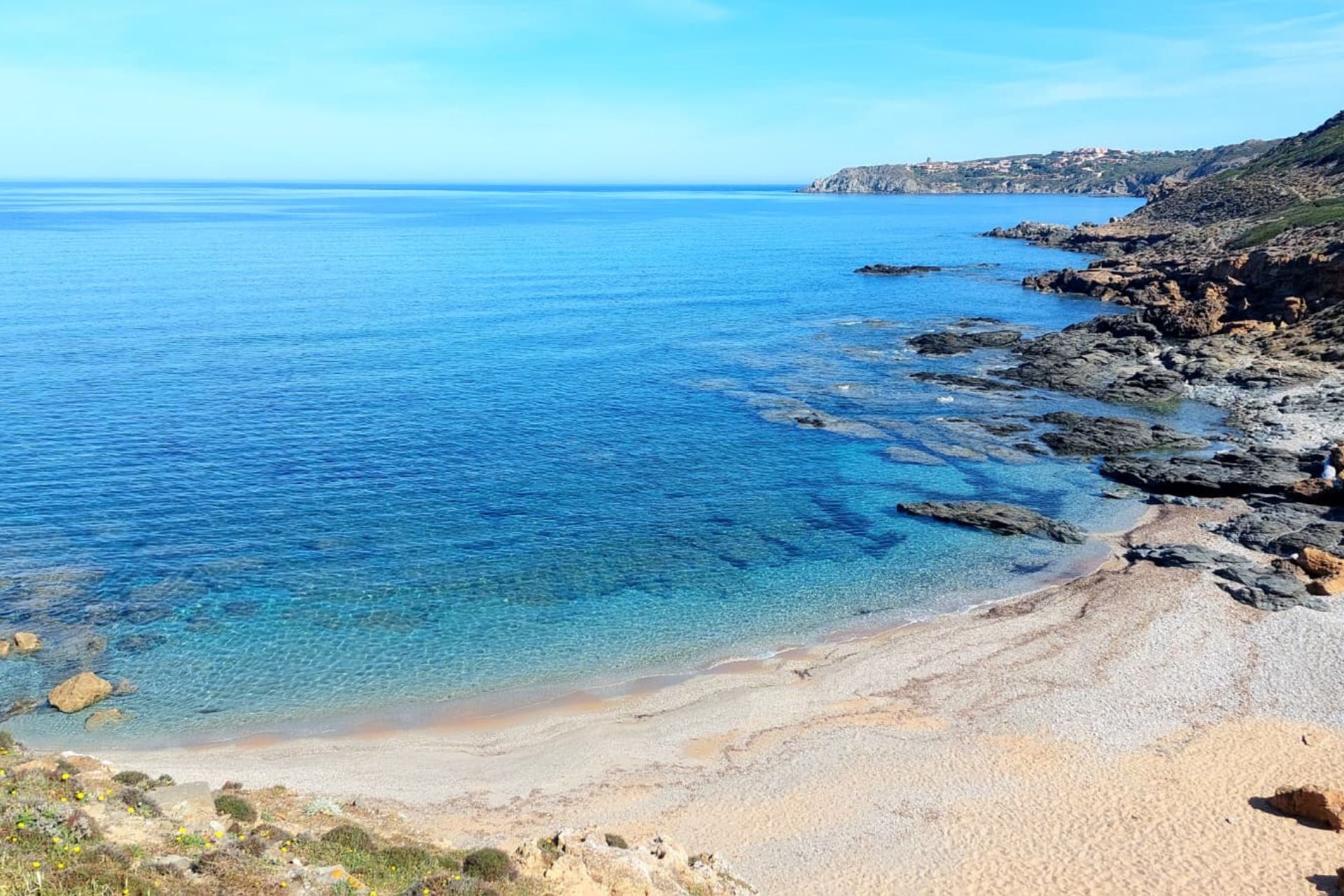 Bild eines Strandes mit blauem Meer und Felsen an der Küste. Im Hintergrund sieht man Hügel und Vegetation
