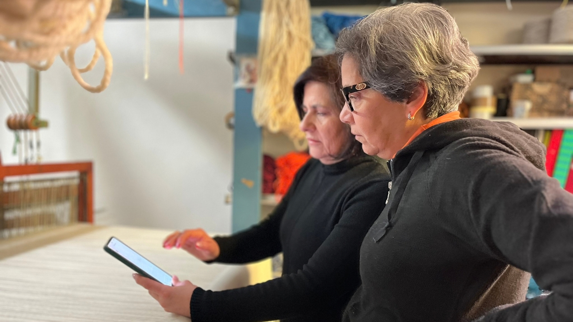 Due donne in un laboratorio di tessitura artigianale, con strumenti e tessuti sullo sfondo