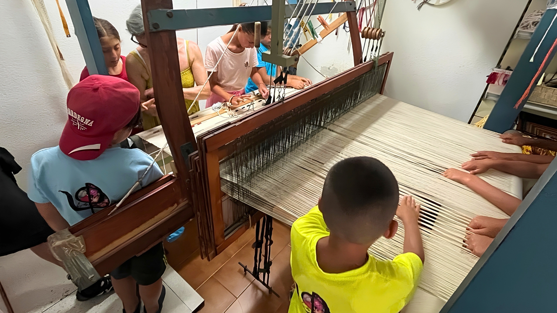 Gruppo di persone, adulti e bambini, intenti a tessere con i telai in un laboratorio artigianale